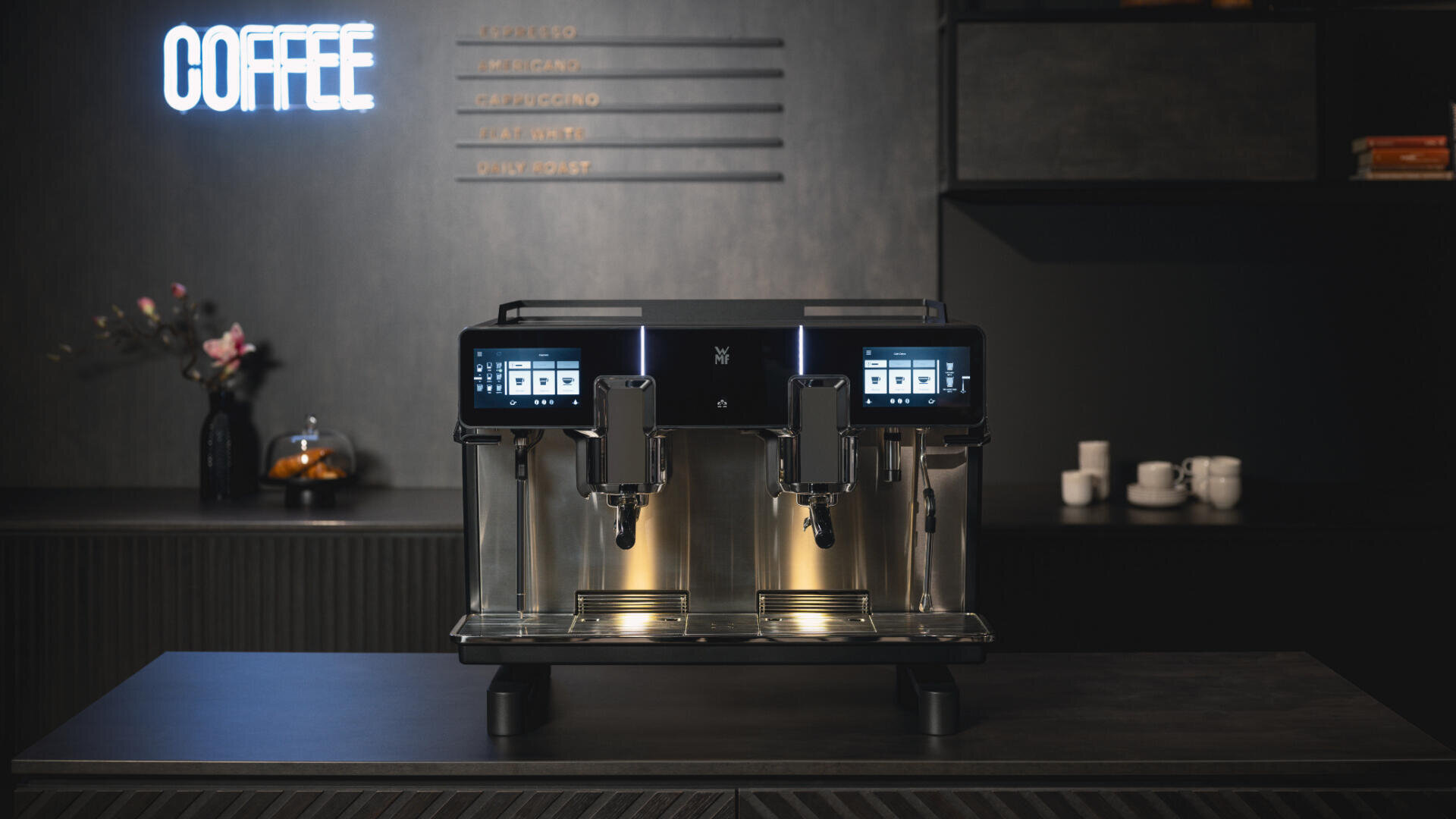 Eine moderne Kaffeemaschine mit zwei Bildschirmen steht auf einer dunklen Theke in einer stilvollen Küche. An der Wand leuchtet ein Neonschild mit der Aufschrift „COFFEE“ und im Hintergrund sind Tassen und Blumen zu sehen.