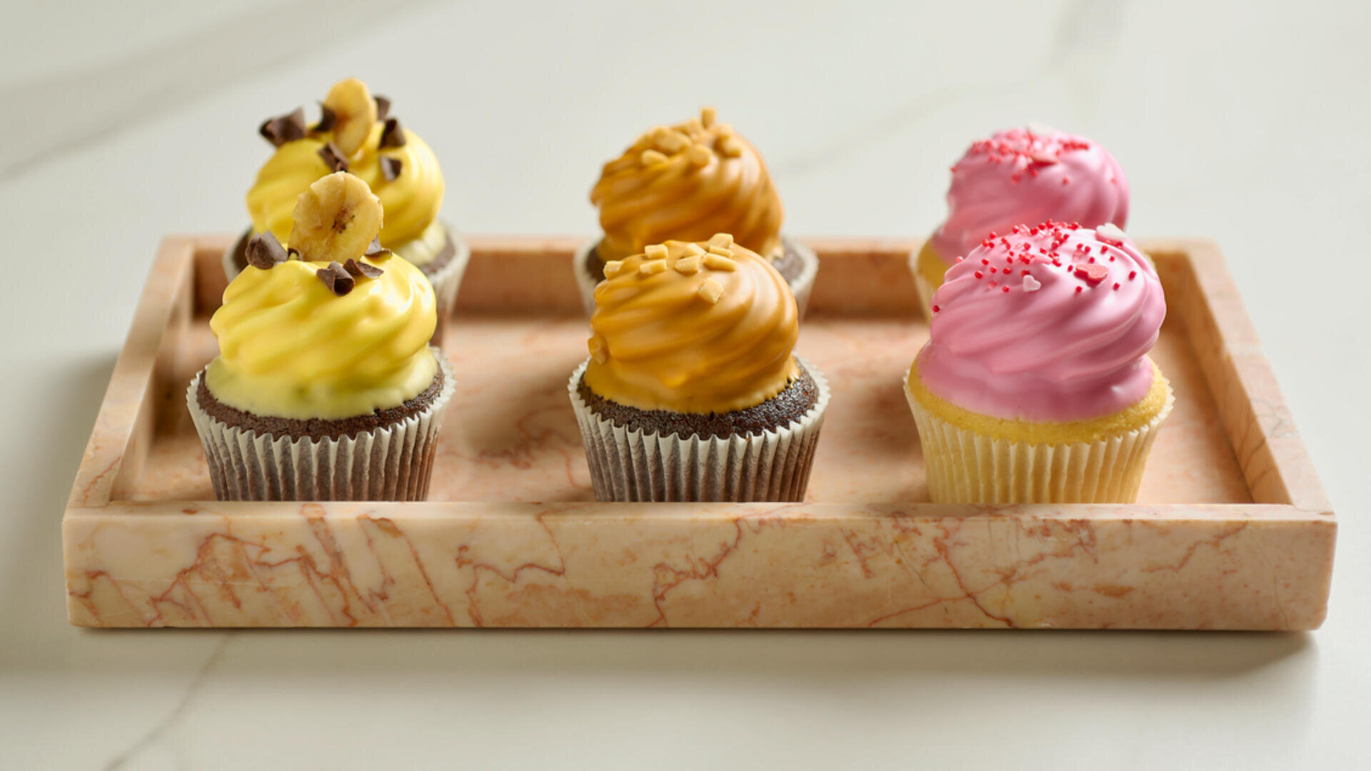 A marble tray holds six cupcakes: two with yellow frosting and banana slices, two with caramel-colored frosting and nut pieces, and two with pink frosting and red sprinkles, arranged neatly in rows.