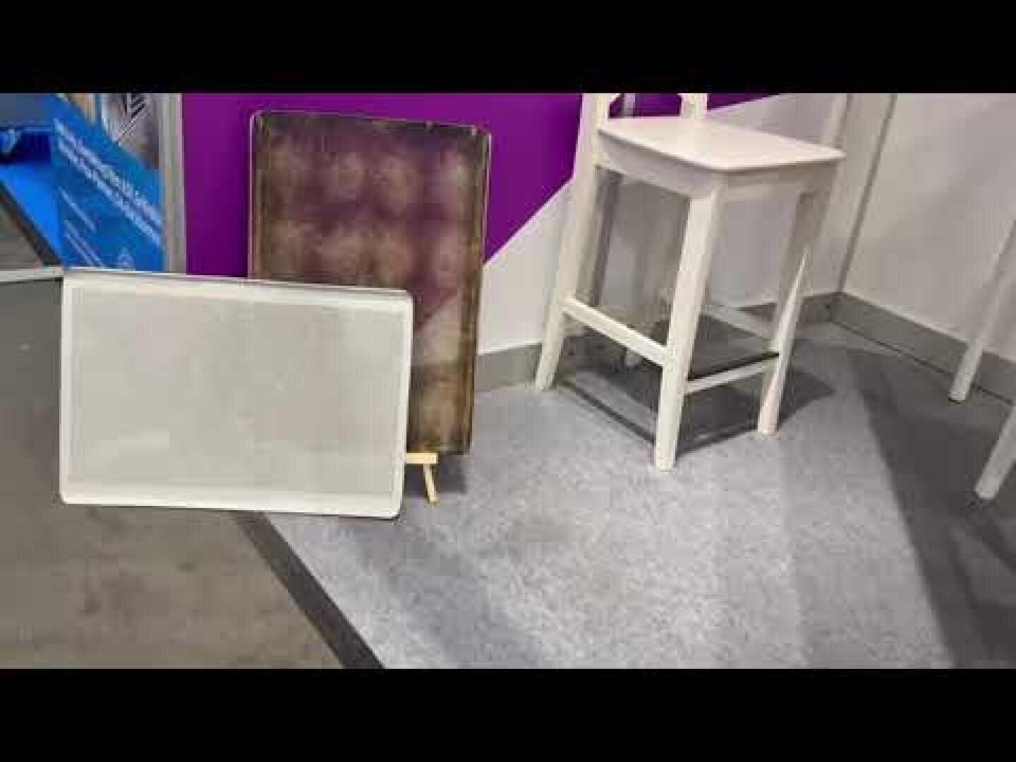 A white chair stands on gray carpet next to two rectangular trays, one metallic and tarnished, the other white and translucent, propped against a purple and white wall at an indoor event or exhibition.