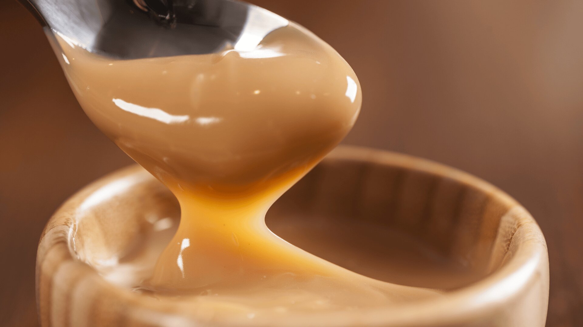 A close-up of a spoon scooping thick, creamy caramel sauce from a wooden bowl, with the sauce stretching smoothly between the spoon and the bowl.
