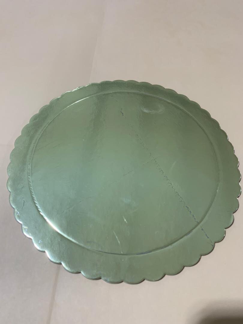 A round, metallic, scalloped-edge cake board sits on a beige surface, reflecting light softly.