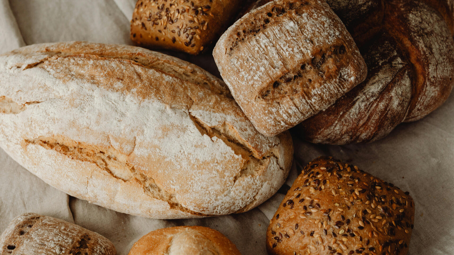 Eine Auswahl an rustikalen Broten und Brötchen mit verschiedenen Formen und gesäten Krusten, die auf einem beigen Tuch angeordnet sind.