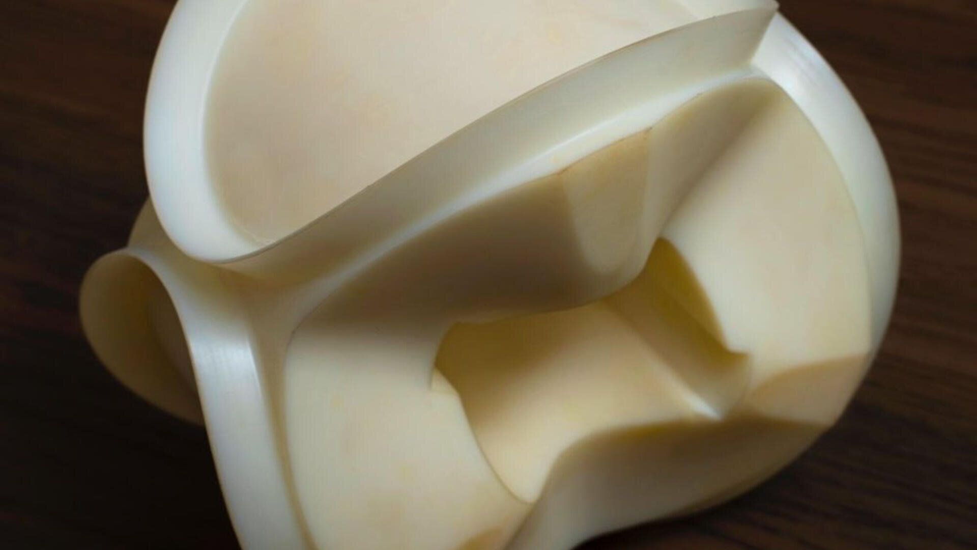 A close-up photo of an abstract, smooth, cream-colored 3D-printed sculpture with curved and intersecting surfaces, placed on a dark wooden surface.