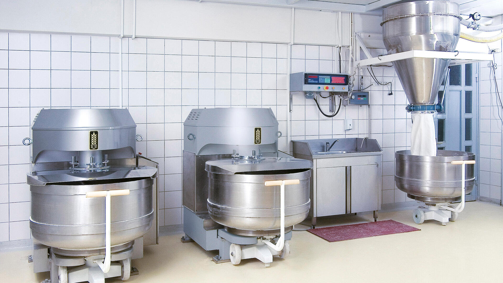 Drei große industrielle Teigknetmaschinen in einer sauberen, gefliesten Bäckereiküche mit Edelstahlspüle, Bedienfeldern und einem Teigtrichter, der die Zutaten in eine der Knetmaschinen einfüllt.