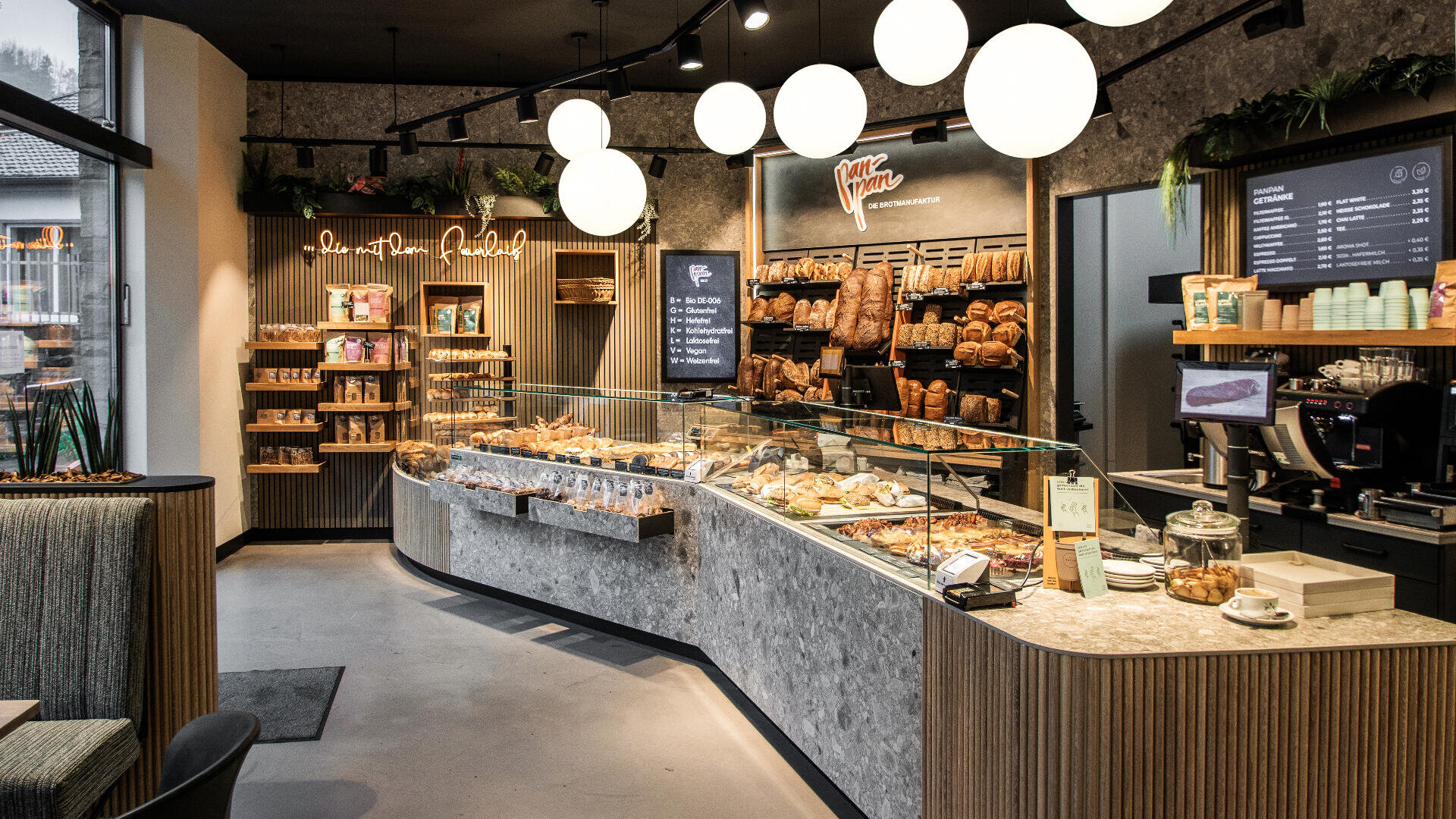 Eine moderne Bäckerei mit einer Glasvitrine, gefüllt mit verschiedenen Backwaren und Brot, Regalen mit Broten im Hintergrund, warmer Beleuchtung und einer digitalen Speisekarte über der Kaffeestation auf der rechten Seite.