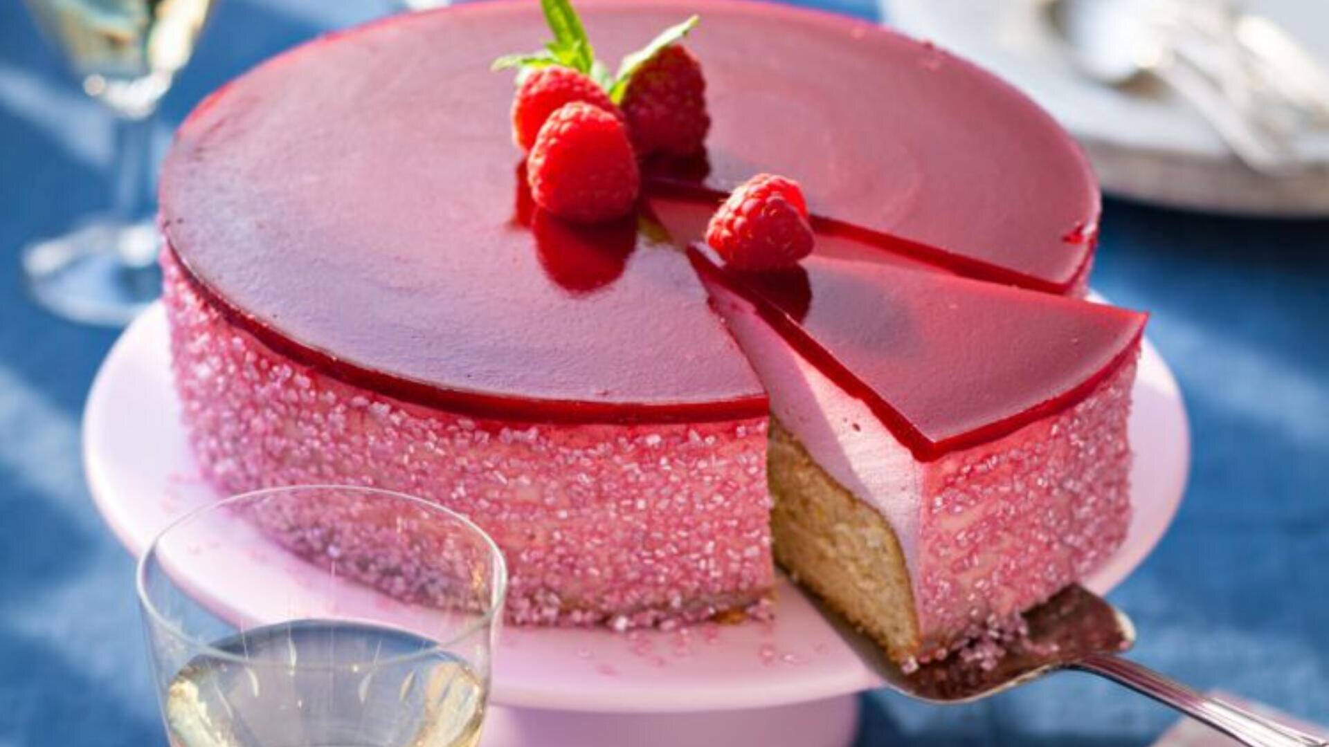 A glossy, red raspberry mousse cake with a slice being served. The cake is decorated with fresh raspberries and a mint sprig on top, and sparkling sugar crystals around the sides. A glass of white wine is nearby.