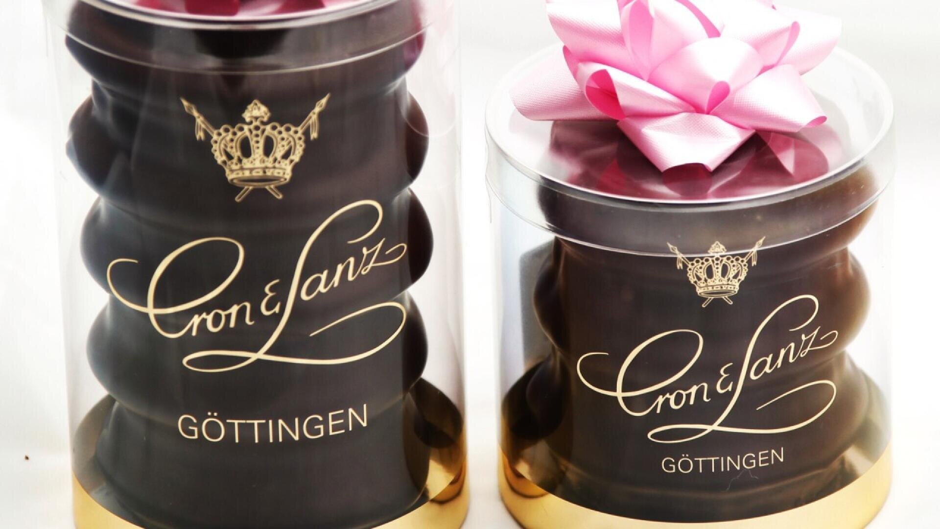 Two clear cylindrical containers with gold bases hold stacked chocolates, each labeled Cron & Lanz Göttingen. Both are topped with a pink ribbon bow. The packaging features a gold crown logo and elegant script.