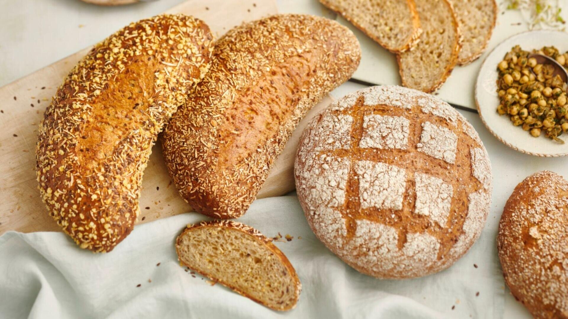 An assortment of fresh whole grain breads, including a round loaf with a crisscross flour pattern and oval loaves topped with seeds, arranged on a wooden board and cloth, with sliced pieces and a bowl of seeds nearby.
