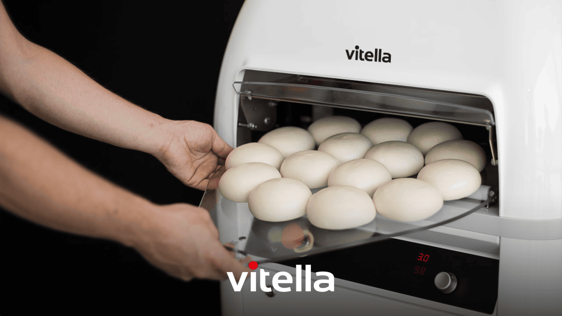 Eine Person legt eine Schale mit runden Teigkugeln in eine weiße, handelsübliche Teigmaschine mit der Aufschrift vitella, deren digitale Bedienelemente auf der Vorderseite sichtbar sind. Das vitella-Logo erscheint deutlich am unteren Rand des Bildes.