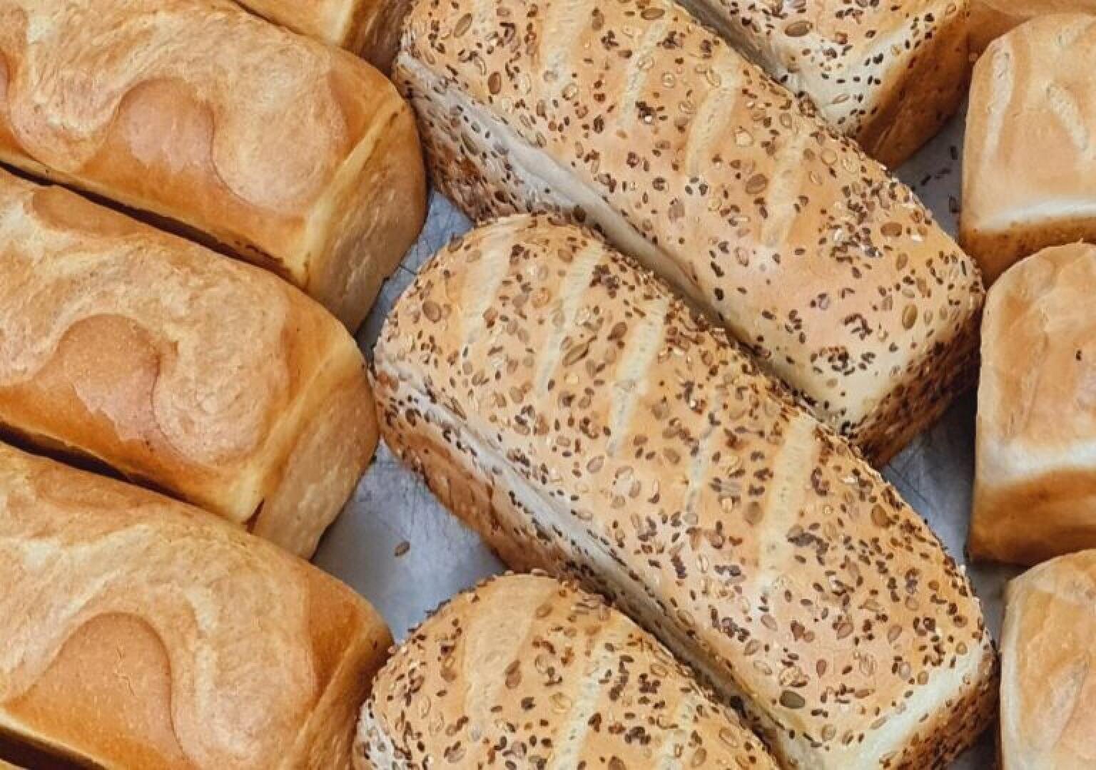 Loaves of bread arranged closely together, including both plain white bread and seeded multigrain bread with a golden-brown crust.