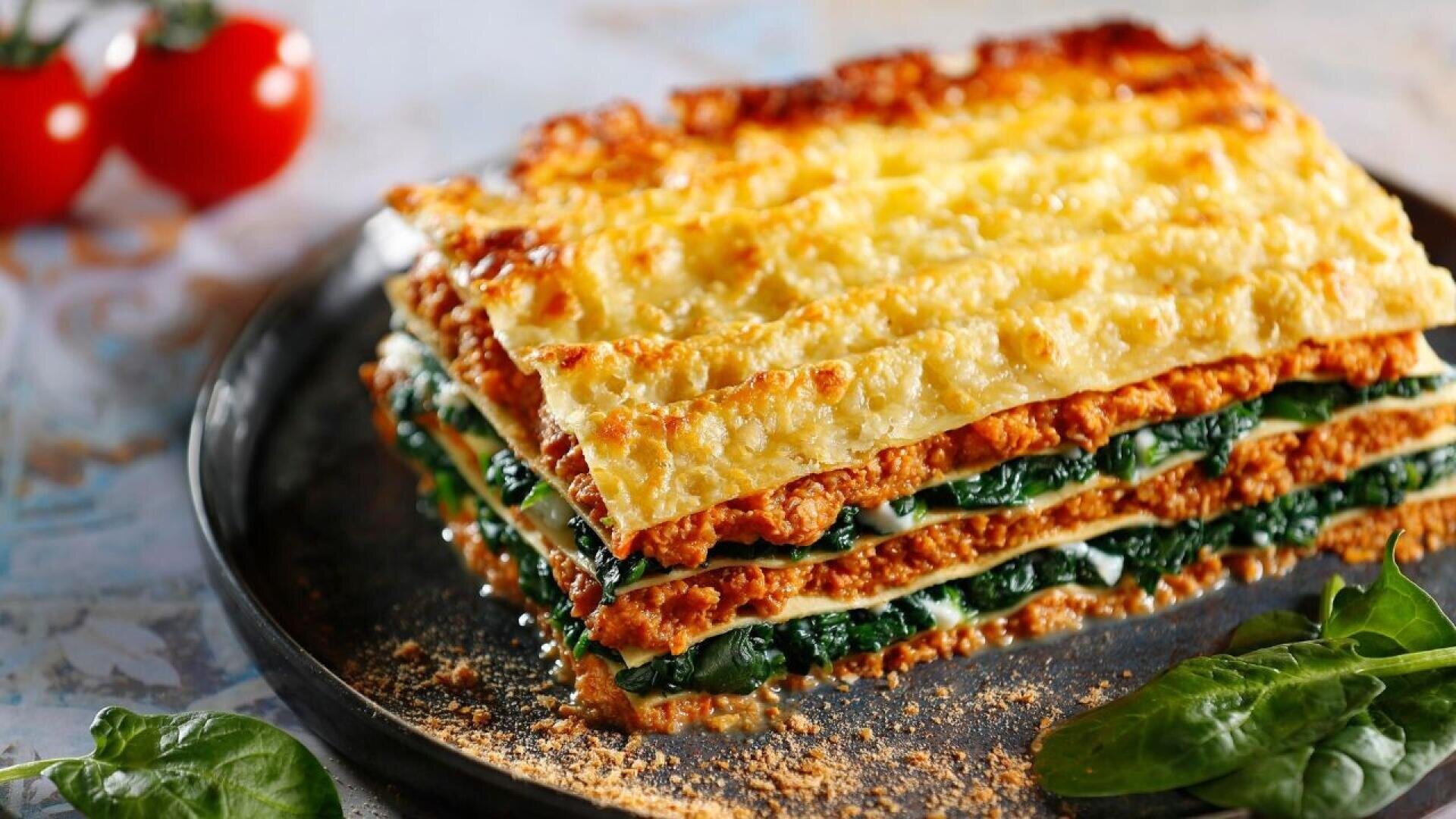 A close-up of a slice of lasagna with golden, crispy cheese on top, layered with spinach, meat sauce, and pasta sheets, served on a dark plate. Fresh spinach leaves and tomatoes are in the background.