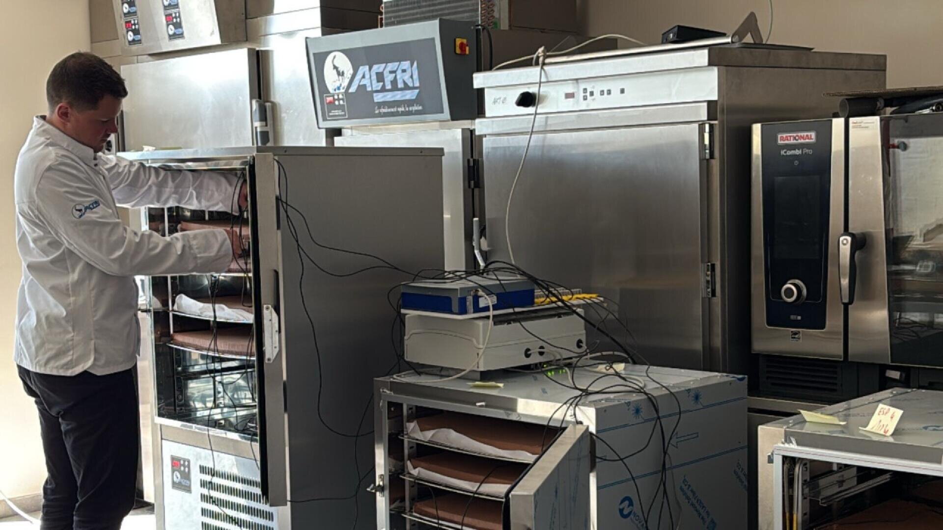 A man in a white lab coat works with ovens and electronic equipment in a commercial kitchen, surrounded by multiple stainless steel appliances and cables.