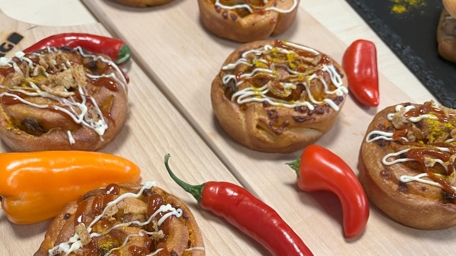 Kleine, runde, herzhafte Gebäckstücke mit Ketchup, Mayonnaise und Röstzwiebeln werden auf Holzbrettern angerichtet, umgeben von roten und orangefarbenen Chilischoten.