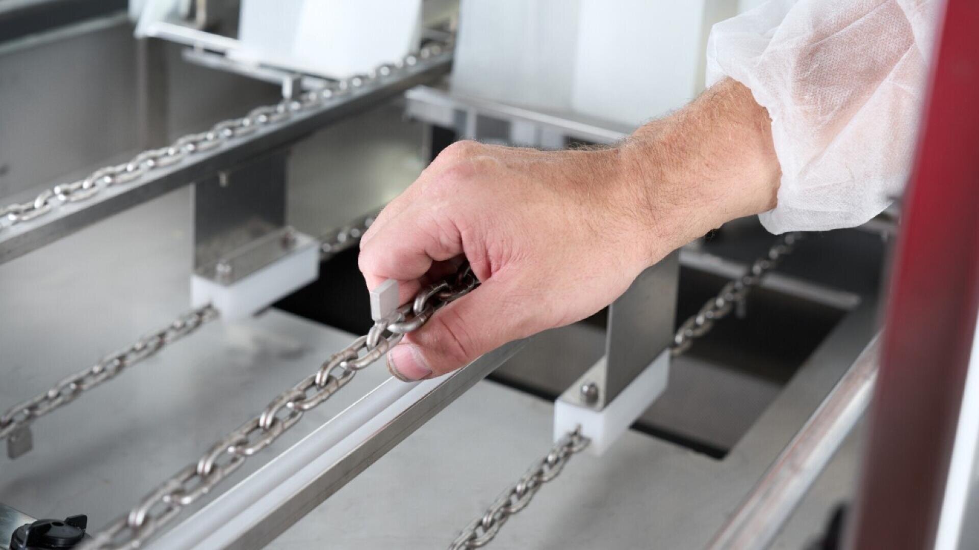 A person wearing a white protective sleeve adjusts a metal chain on a machine, likely in an industrial or manufacturing setting.