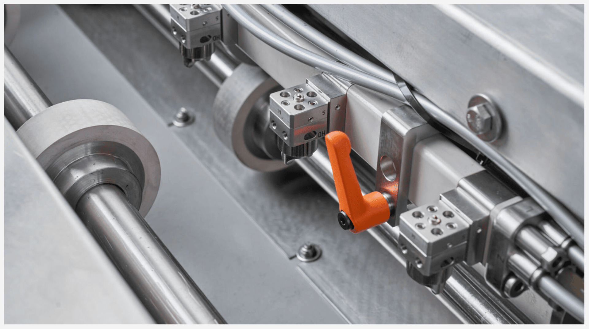 Nahaufnahme einer Industriemaschine aus Metall mit Rollen, Sensoren und einem orangefarbenen Hebelgriff, der verschiedene Kabel und Metallteile aufweist.