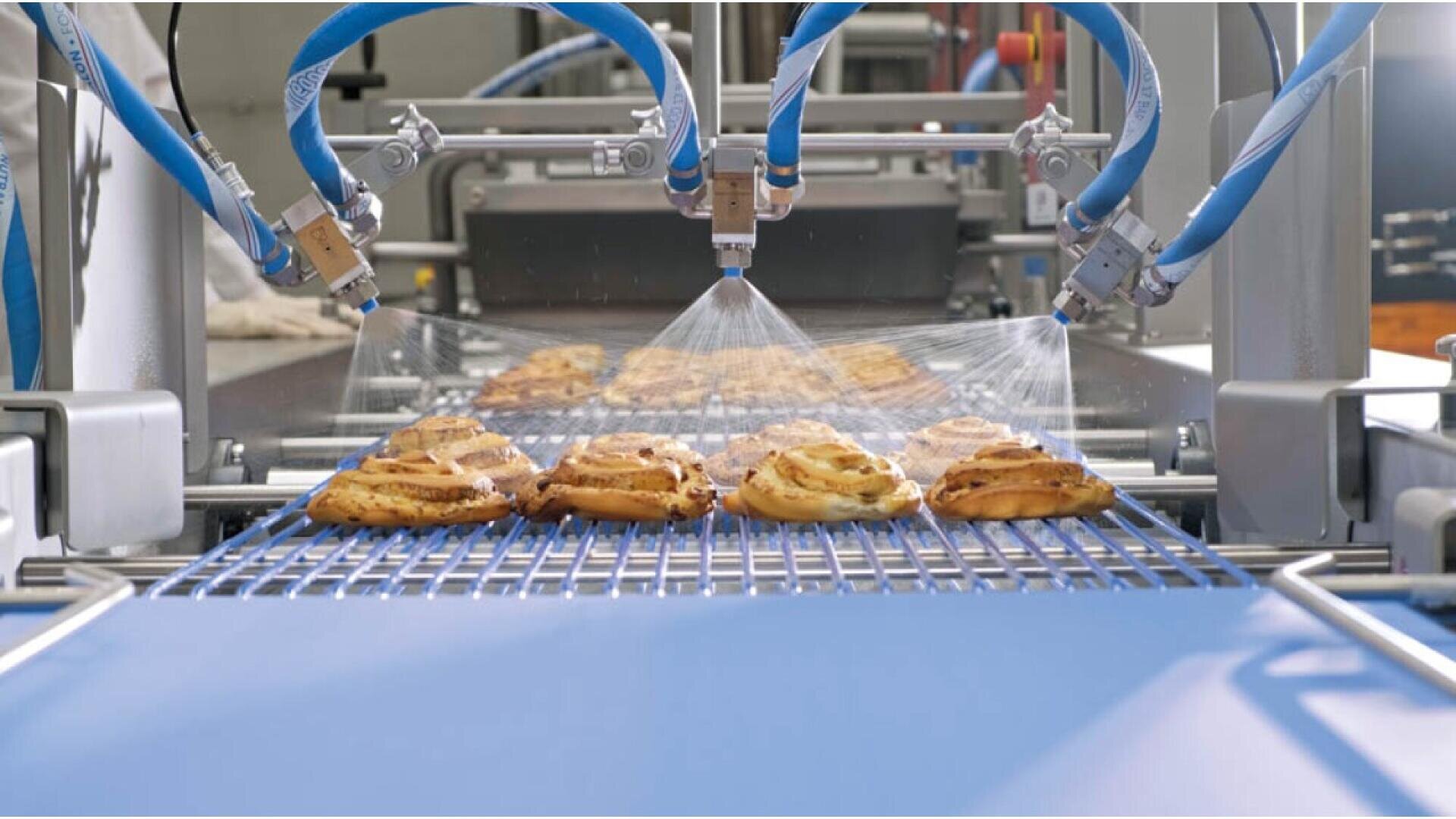 Nahaufnahme von Gebäck auf einem Förderband, das in einer Lebensmittelfabrik von automatisierten Düsen besprüht wird. Die Maschine trägt eine Beschichtung oder Glasur auf das Gebäck auf, während es sich entlang der Produktionslinie bewegt.