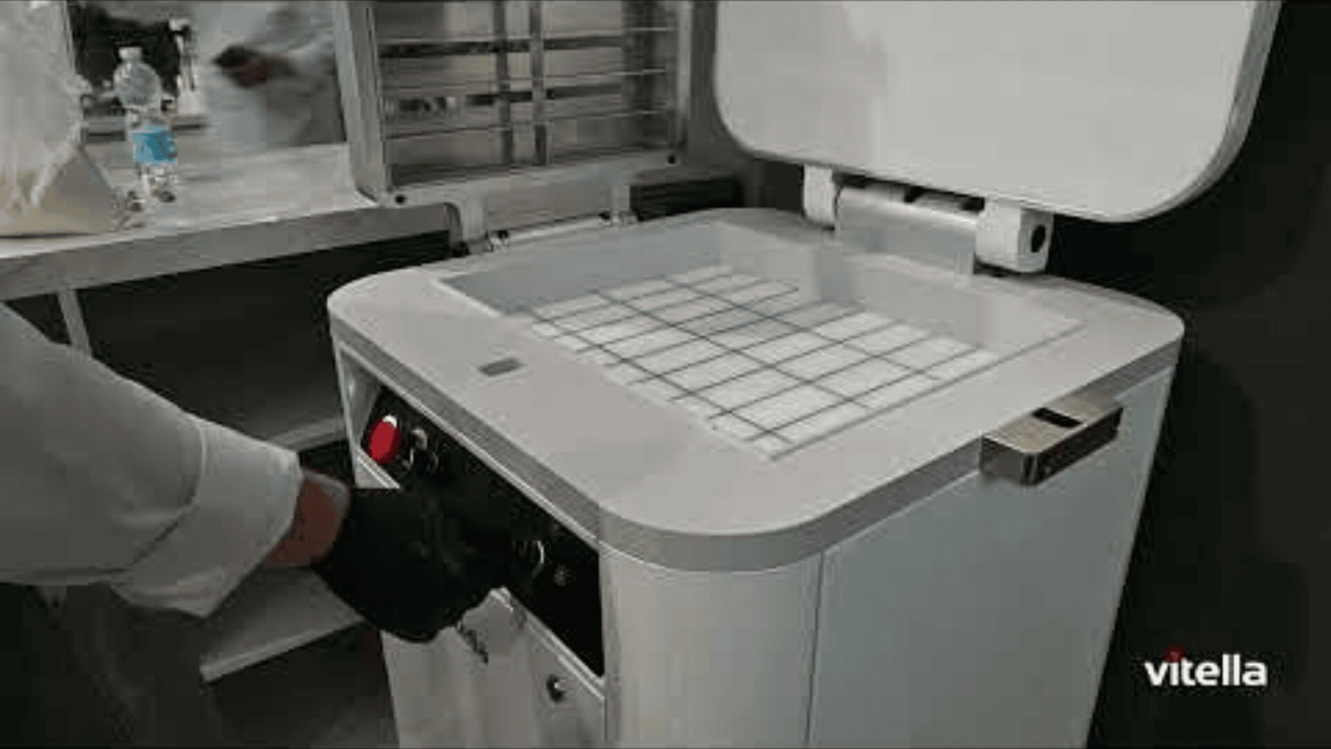 A person wearing a black glove is operating a white machine with a grid surface and an open lid in a laboratory or kitchen. The vitella logo can be seen in the bottom right-hand corner.