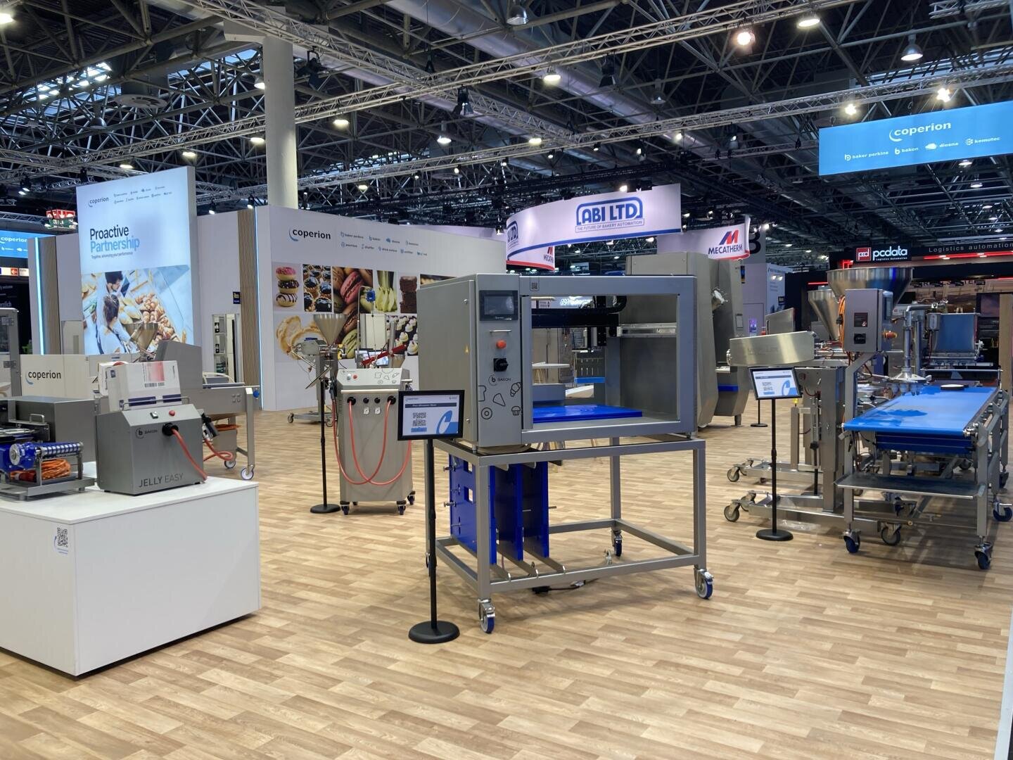 A trade show booth displays various industrial food processing machines on a wooden floor, with bright overhead lighting and company banners, including one for Coperion, in the background.