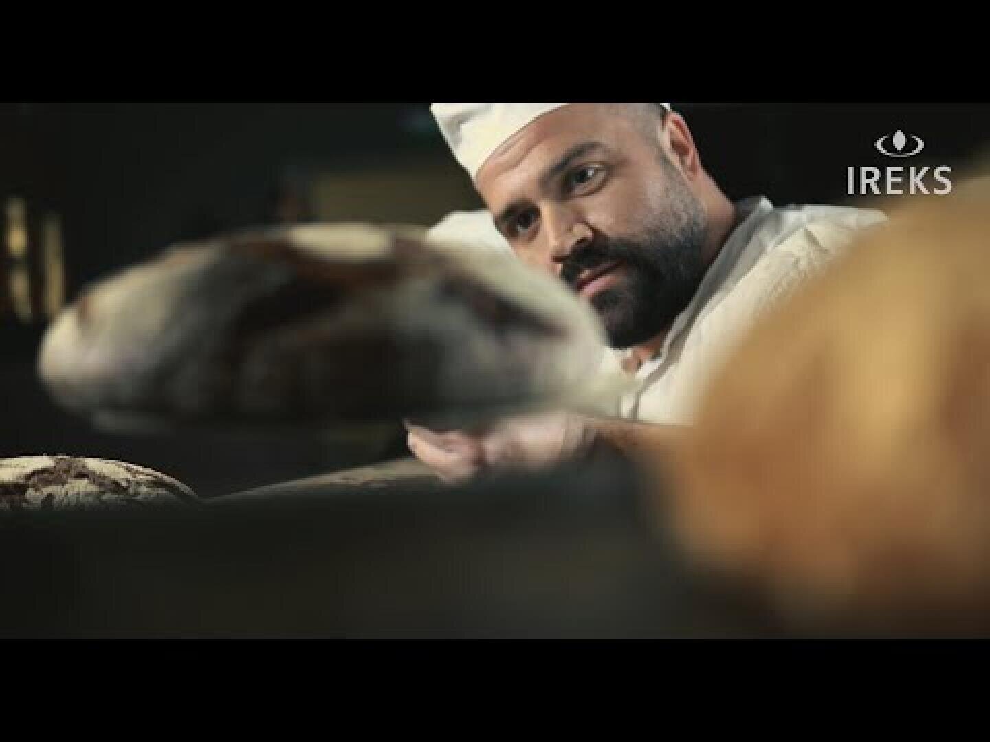 A baker in a white cap and uniform examines fresh loaves of bread. The IREKS logo is visible in the upper right corner. The focus is on the bread in the foreground and the bakers attentive facial expression.