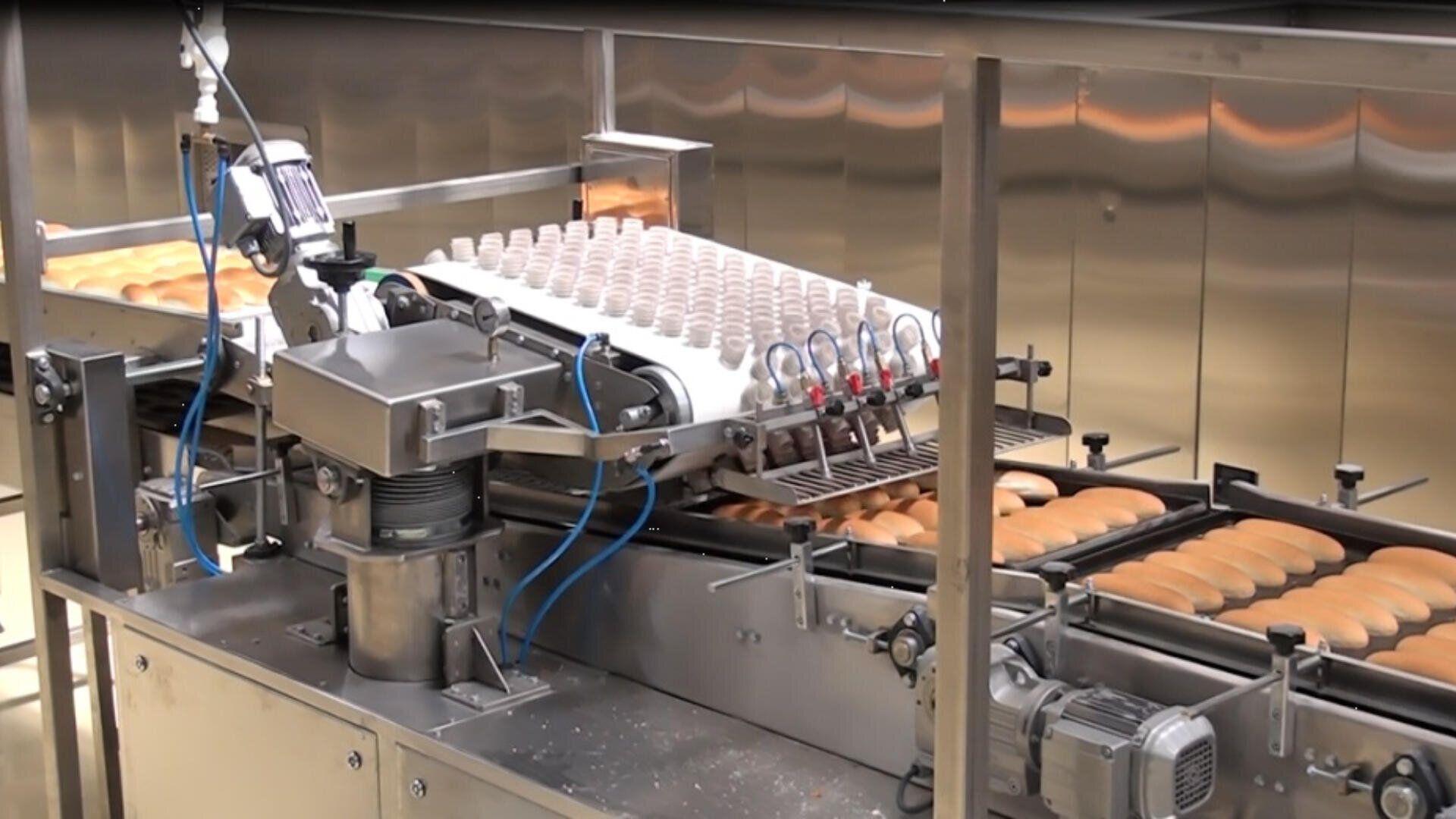 A machine in a factory places cream filling onto rows of pastries or buns moving along a conveyor belt, automating the filling process in a commercial bakery setting.