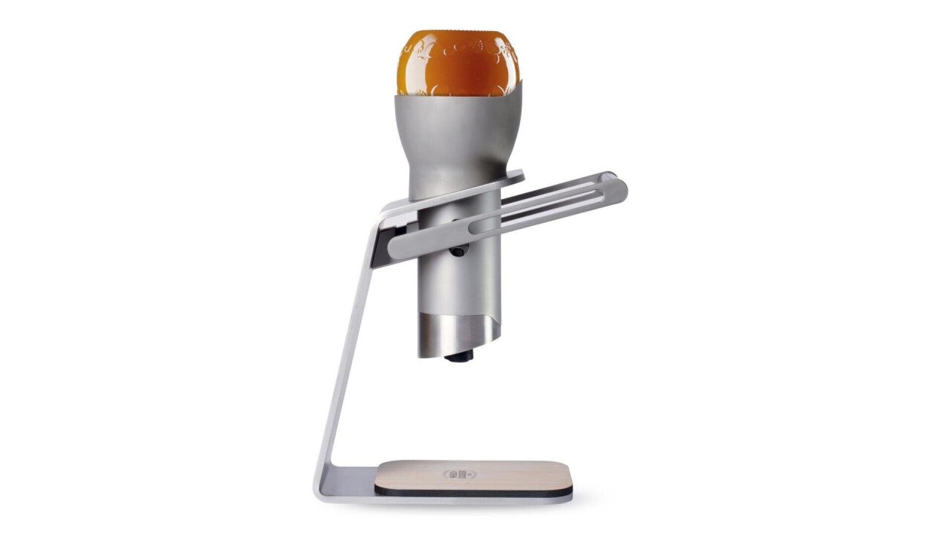 A modern beer dispenser with a sleek, metallic design holds an upside-down beer bottle. The device stands on a minimalist base with a wooden accent.