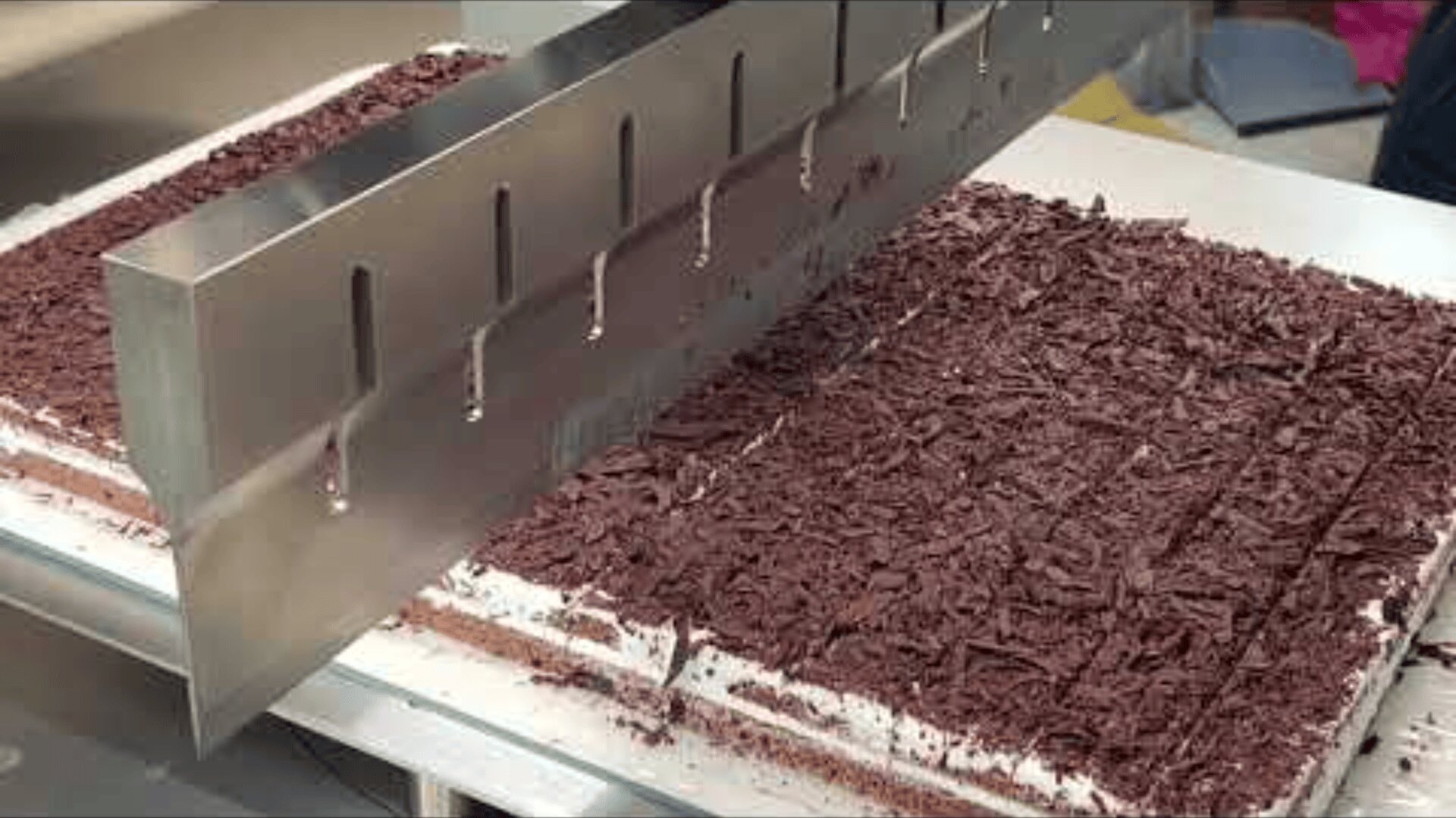 A large metal knife cuts a rectangular cake with chocolate shavings into even pieces on a metal surface.
