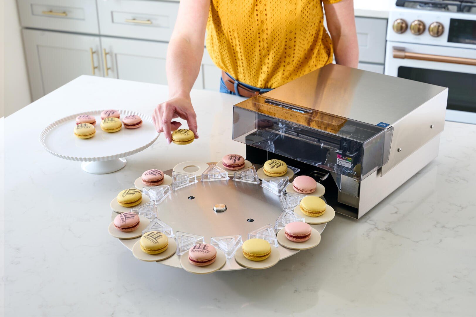 A person uses a machine to print designs onto macarons, placing decorated and plain macarons on a white kitchen counter next to a plate with more macarons.
