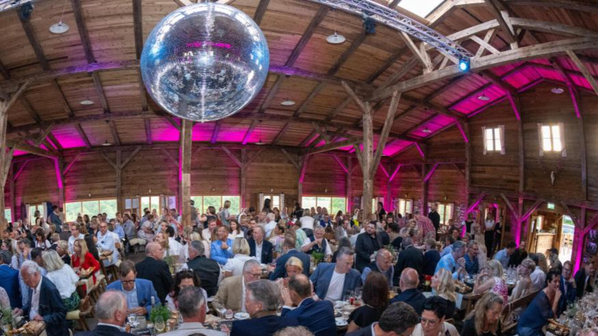 Eine große Gruppe formell gekleideter Menschen sitzt an Tischen in einer rustikalen Holzhalle mit hoher Decke, violetter Beleuchtung und einer riesigen Discokugel darüber.
