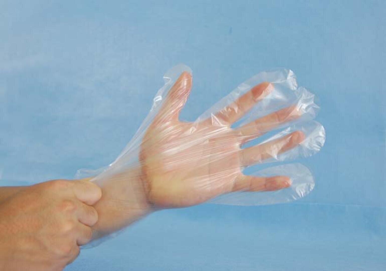 A person puts on a transparent disposable plastic glove against a light blue background.