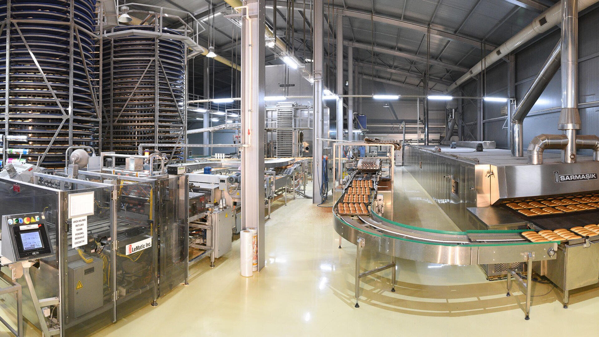 A modern industrial bakery with automated machines, conveyor belts transporting baked goods, and towering racks in a spacious, well-lit factory setting.