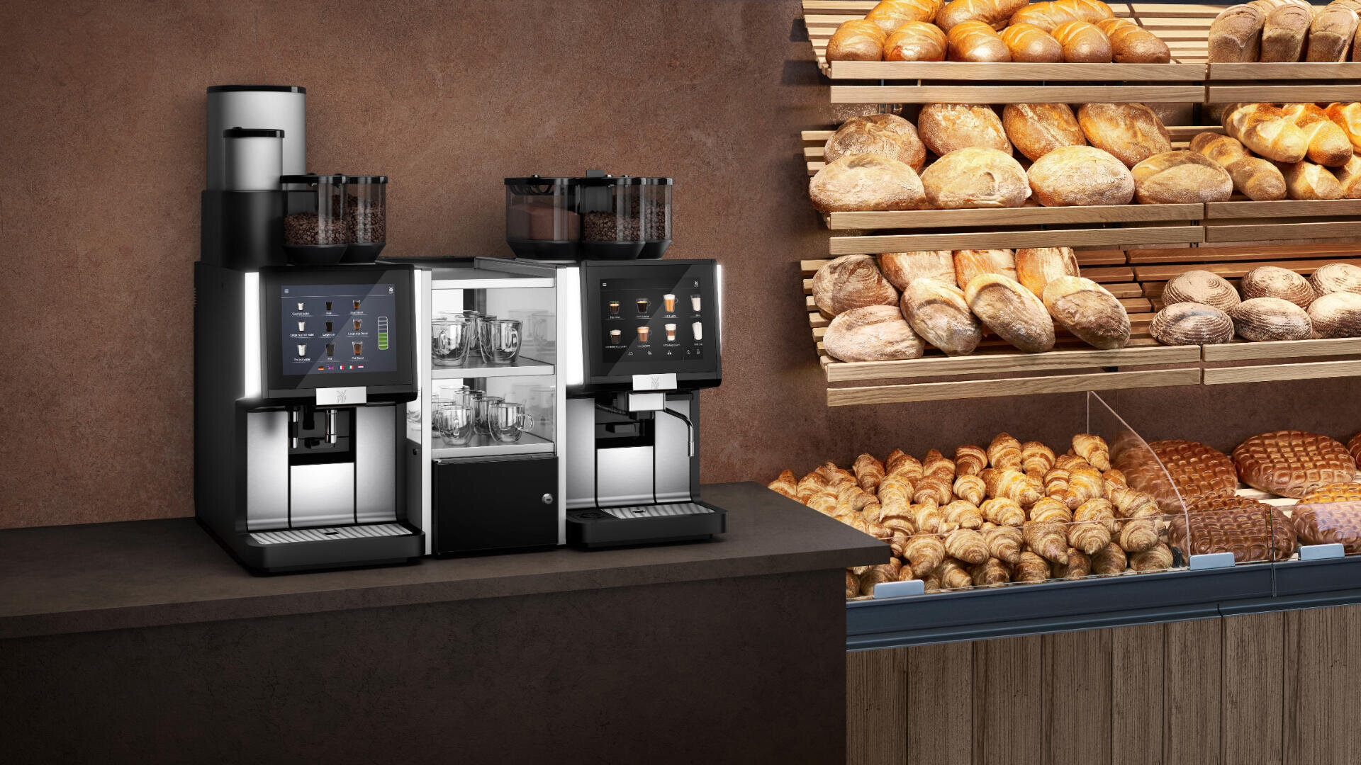 Zwei moderne Kaffeemaschinen stehen auf einer Theke neben Regalen voller frischer Brot- und Gebäcksorten in einer Bäckereiumgebung mit braunem Wandhintergrund.