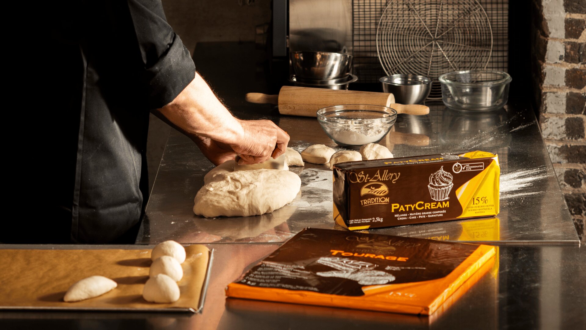 A person kneads dough on a metal countertop in a kitchen, with baking ingredients, tools, and packaged butter nearby. Dough portions are ready on a baking tray. The scene is warmly lit.
