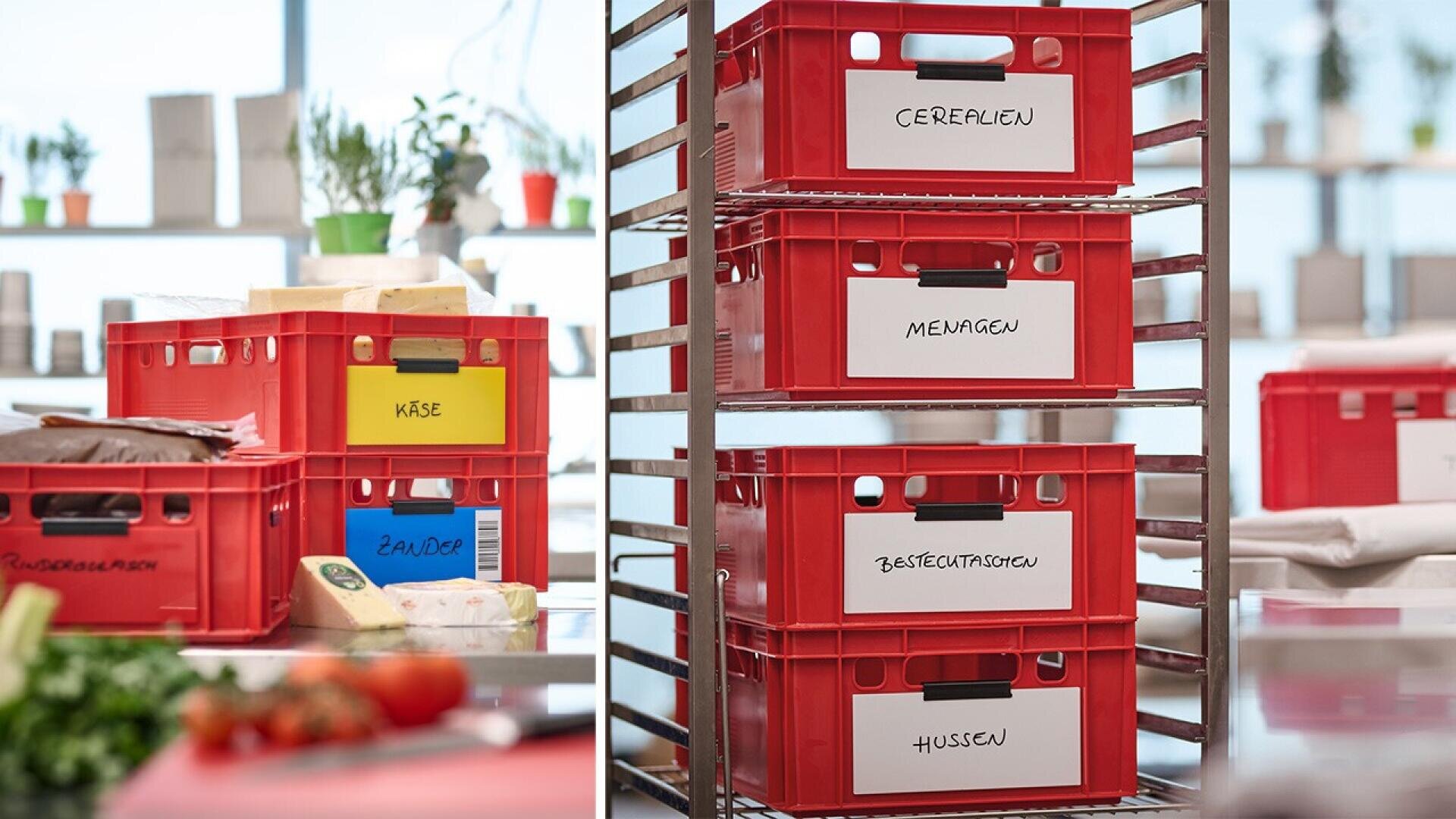 Red plastic crates with white labels, containing food items, are stacked on metal racks in a bright kitchen. The labels are in German, including words like Käse, Cerealien, and Hülsen. Some fresh produce is visible nearby.