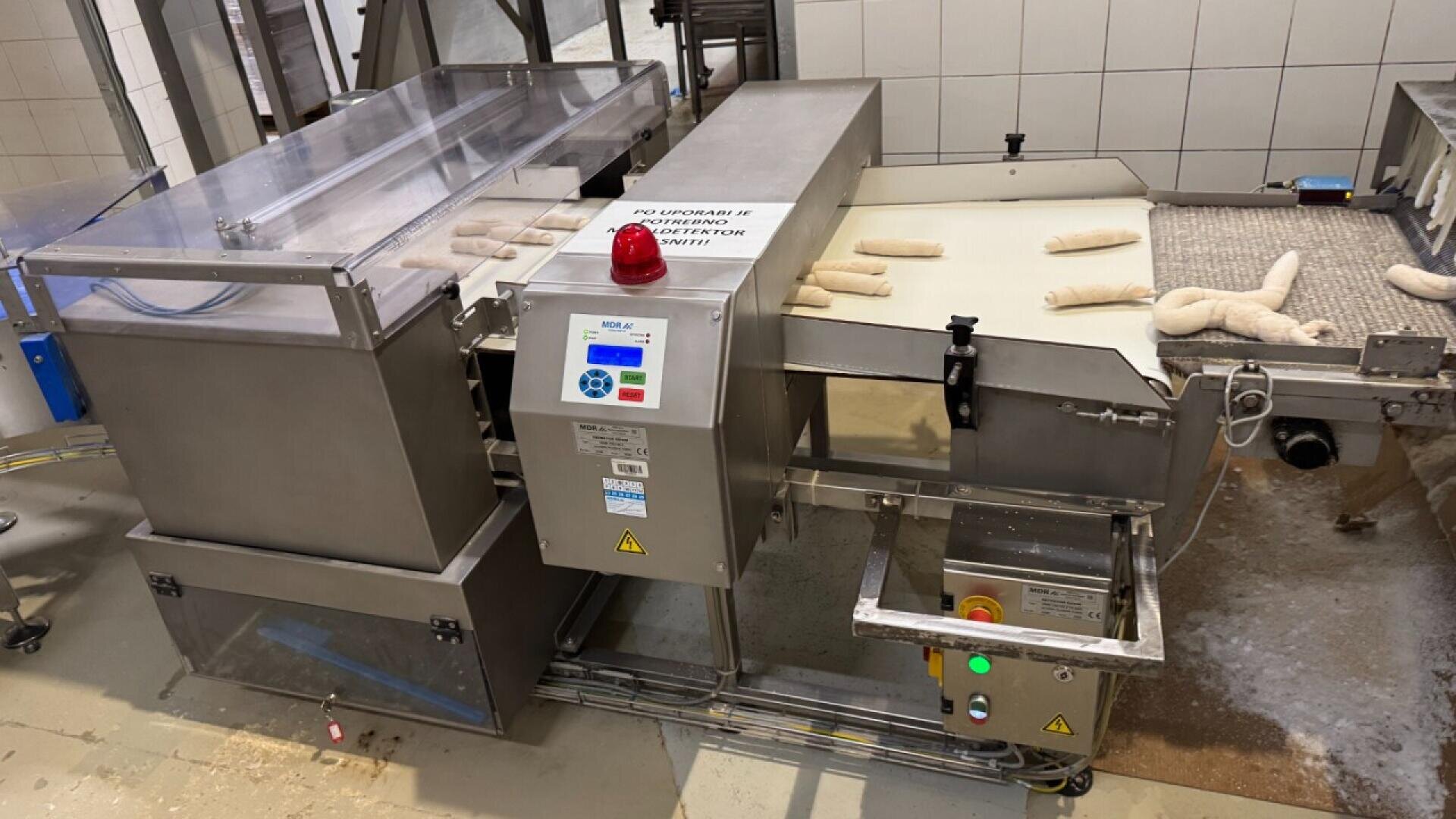 Eine Brotproduktionsmaschine formt auf einem Förderband in einer Bäckerei den Rohteig zu Baguetteformen. Der Bereich ist sauber und industriell, mit gefliesten Wänden und Geräten aus Edelstahl.