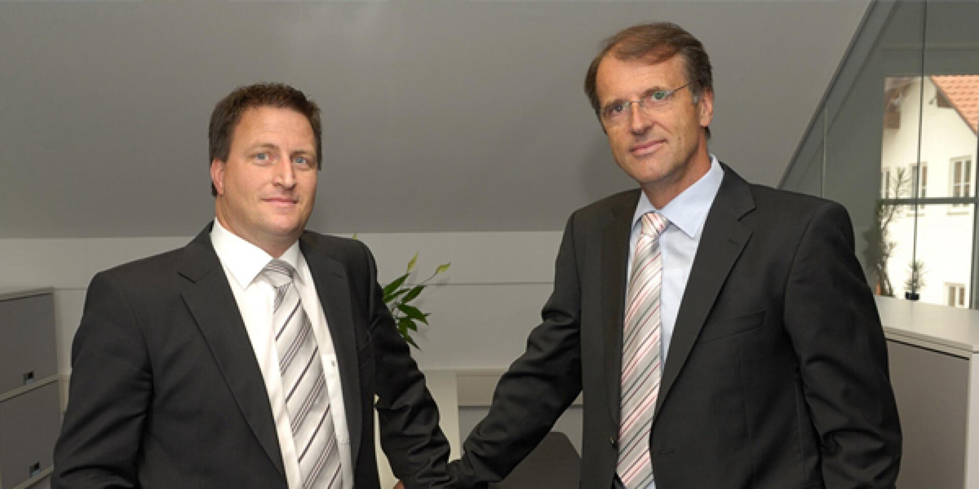 Two men in business suits stand in an office, both looking at the camera. One man has his hand resting on a desk. There is a plant and file cabinets in the background.