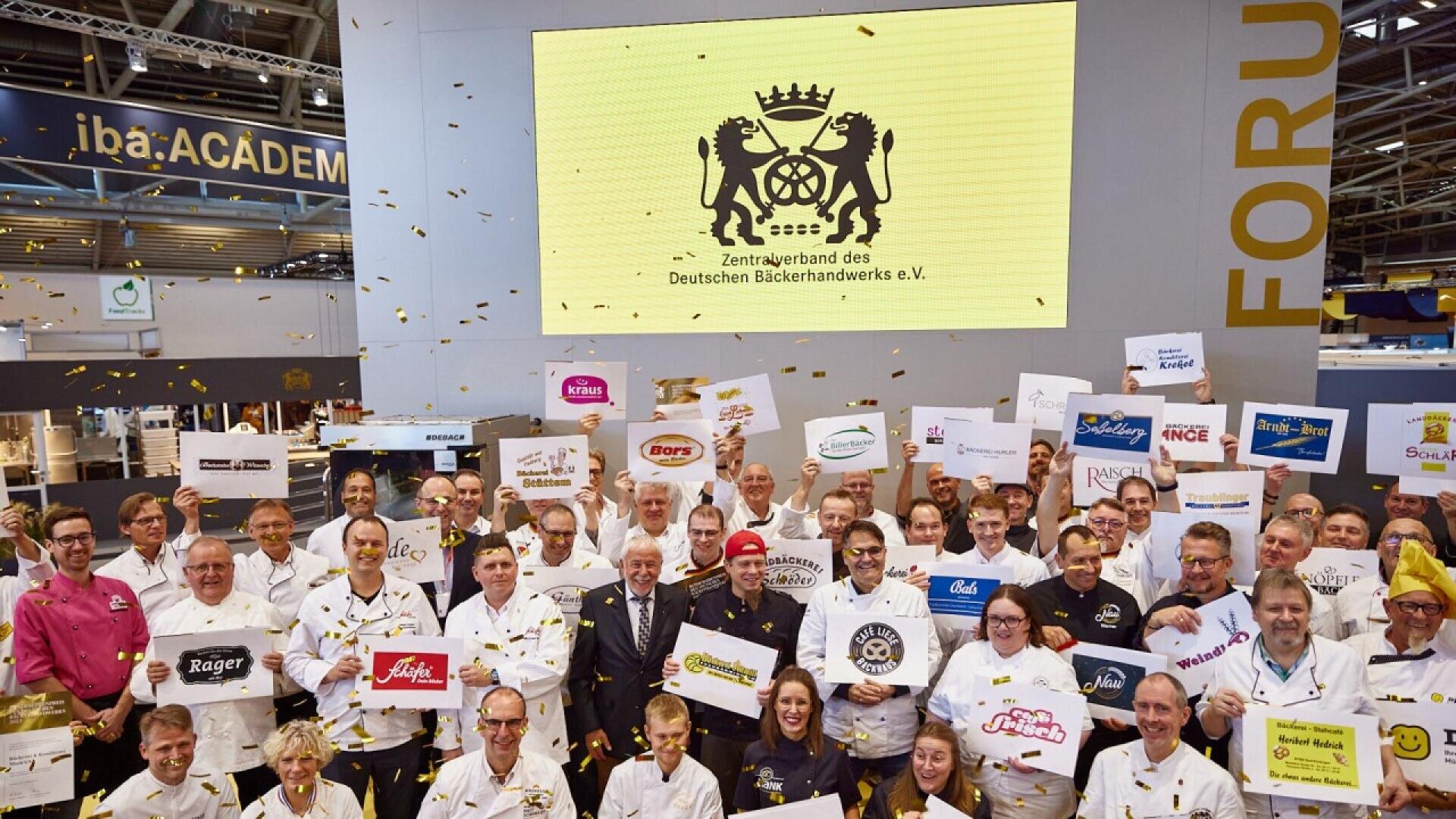 A large group of people in white uniforms stand smiling, holding bakery brand signs under a screen with the logo of the Zentralverband des Deutschen Bäckerhandwerks, surrounded by gold confetti at a bakery event.