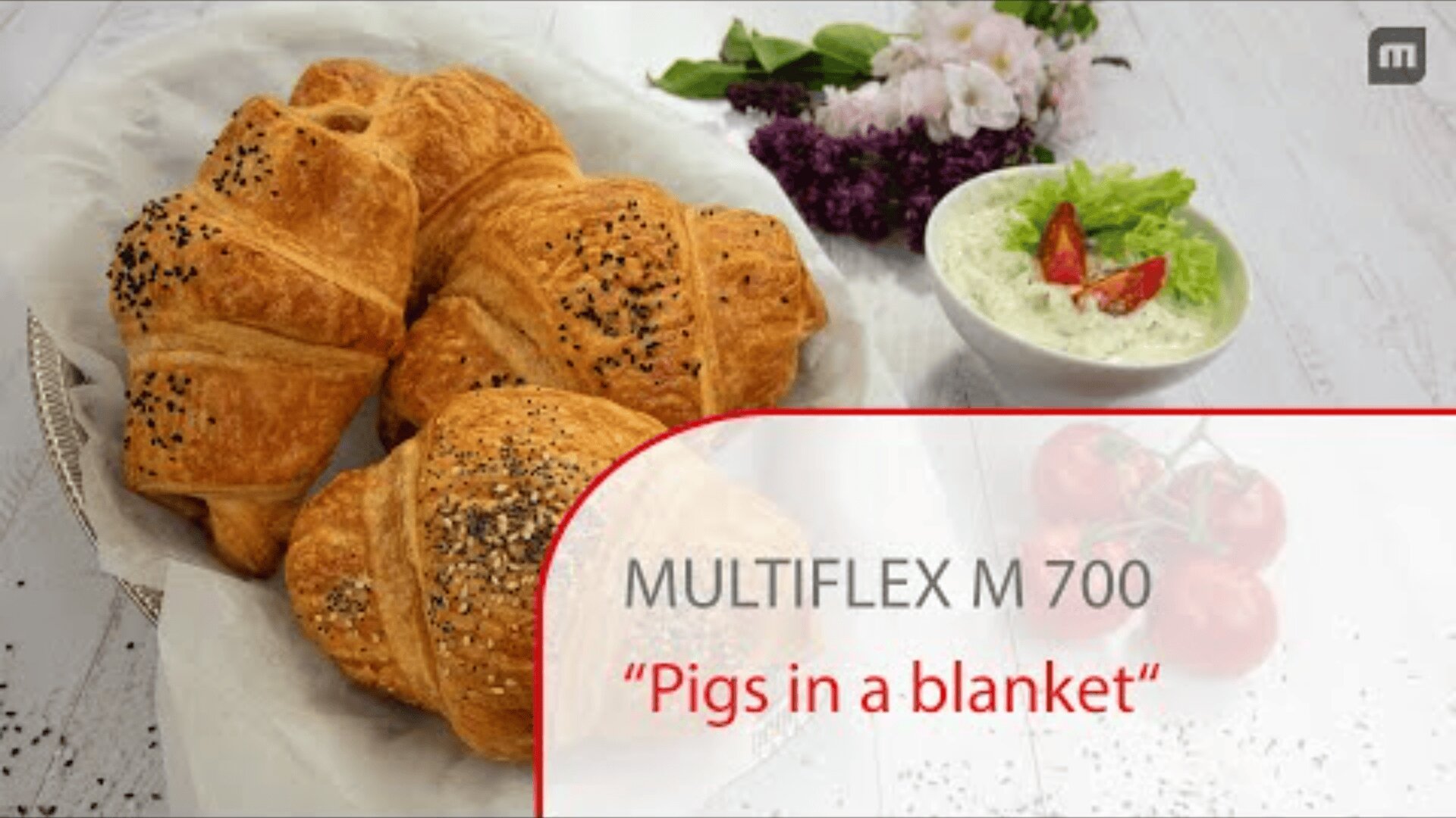 A plate of golden, flaky croissant-style pigs in a blanket with sesame and poppy seeds, beside a bowl of salad with lettuce and tomato, and purple flowers in the background. Text reads: MULTIFLEX M 700 Pigs in a blanket.
