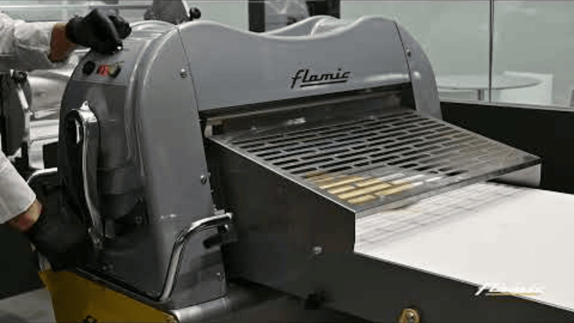 One person operates a large industrial dough sheeter called Flamic, where the dough is fed through rollers onto a conveyor belt.