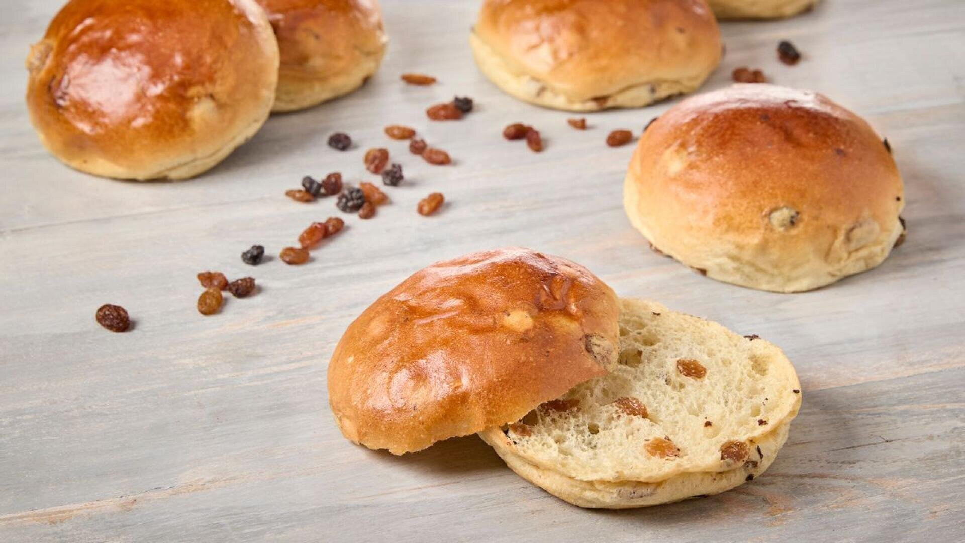Mehrere glänzende, goldbraune Rosinenbrötchen liegen auf einer hellen Holzoberfläche. Ein Brötchen ist aufgeschnitten und zeigt seine weiche, luftige Textur sowie die verstreuten Rosinen. Lose Rosinen sind daneben verstreut.