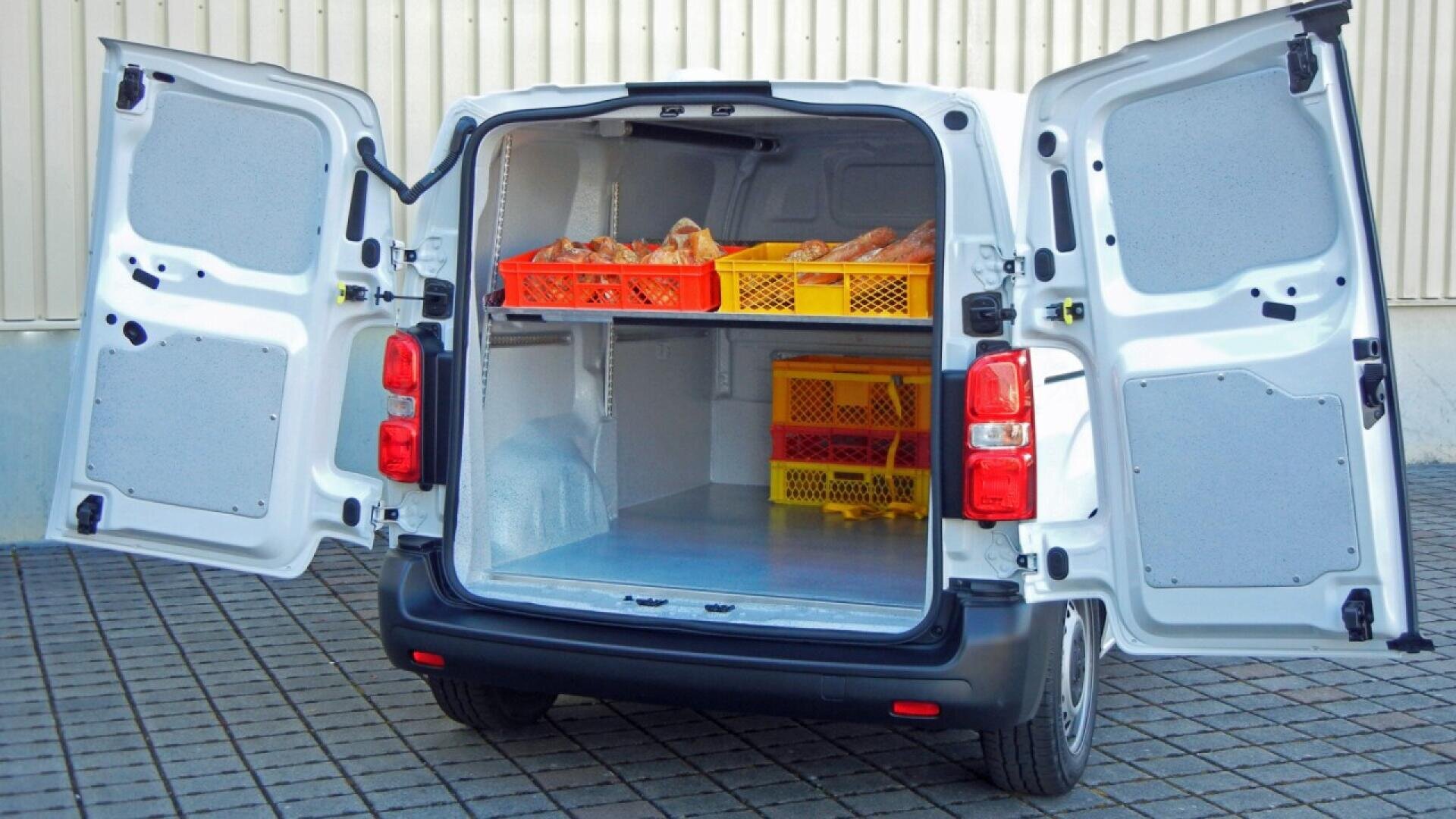 A white delivery van with both rear doors open reveals crates of bread and baked goods stacked on shelves inside. The van is parked on a patterned brick surface near a beige building.