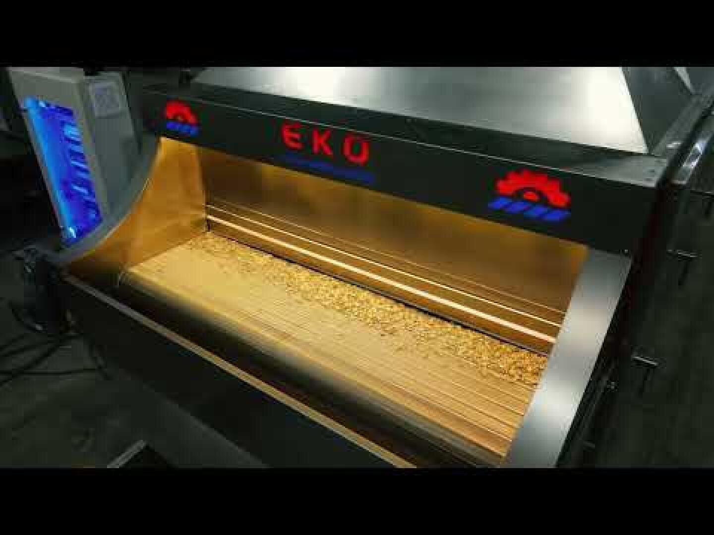A commercial roasting machine labeled EKO is filled with a layer of roasted coffee beans. The machine features metallic surfaces and indicator lights on the front.