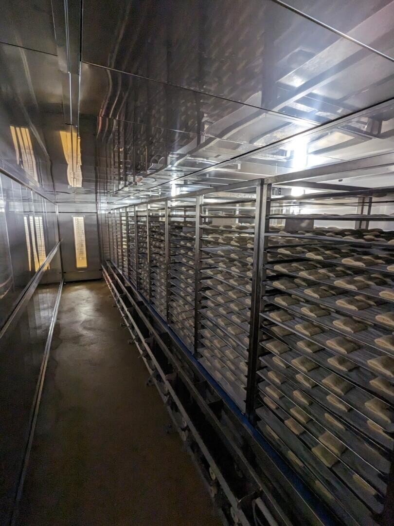 Ein langer, schmaler Industrieraum mit Metallregalen, auf denen mehrere Tabletts mit Teigstücken stehen, möglicherweise in einer Bäckerei oder einem Lebensmittelverarbeitungsbetrieb, unter hellem Leuchtstoffröhrenlicht.