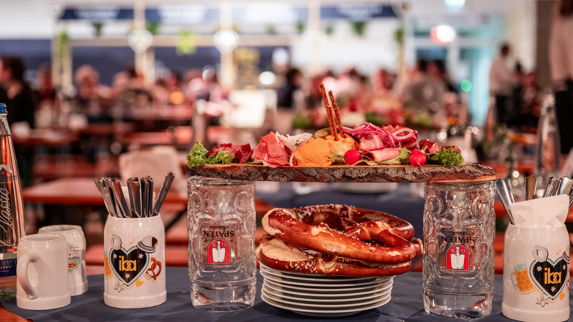 Ein Tisch mit großen Brezeln, Aufschnitt, Käse, Radieschen und Bierkrügen mit Oktoberfestmotiven in einer lebhaften, verschwommenen Feststimmung.