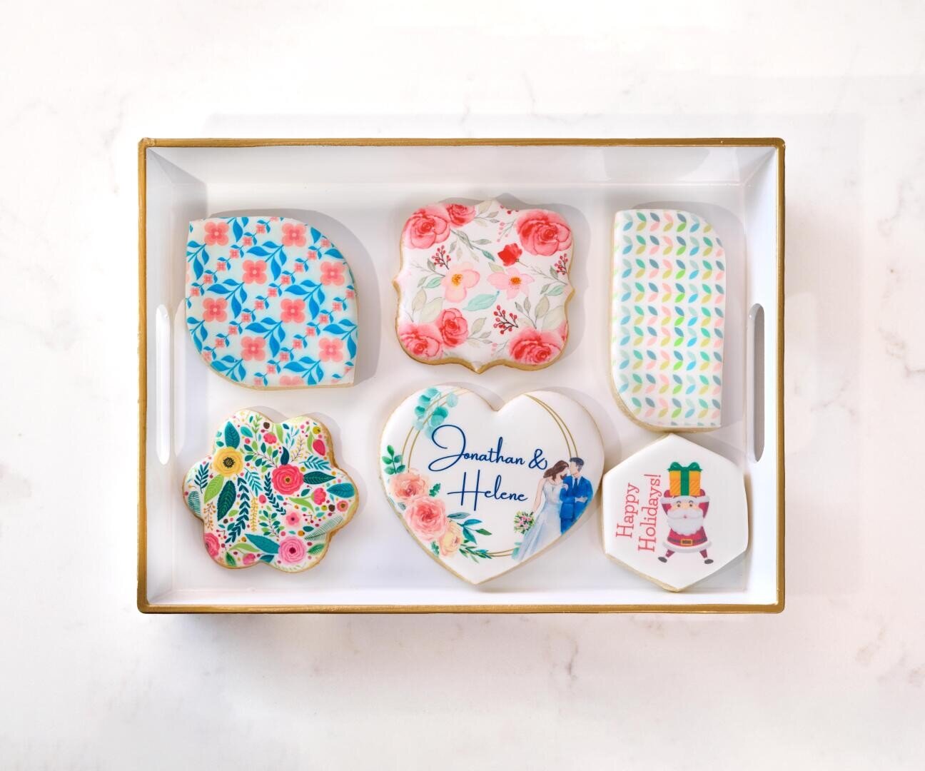 A box with six decorated cookies, each with colorful floral or geometric patterns; one heart-shaped cookie says Jonathan & Helene and a round cookie reads Happy Holi-day! with a festive design.