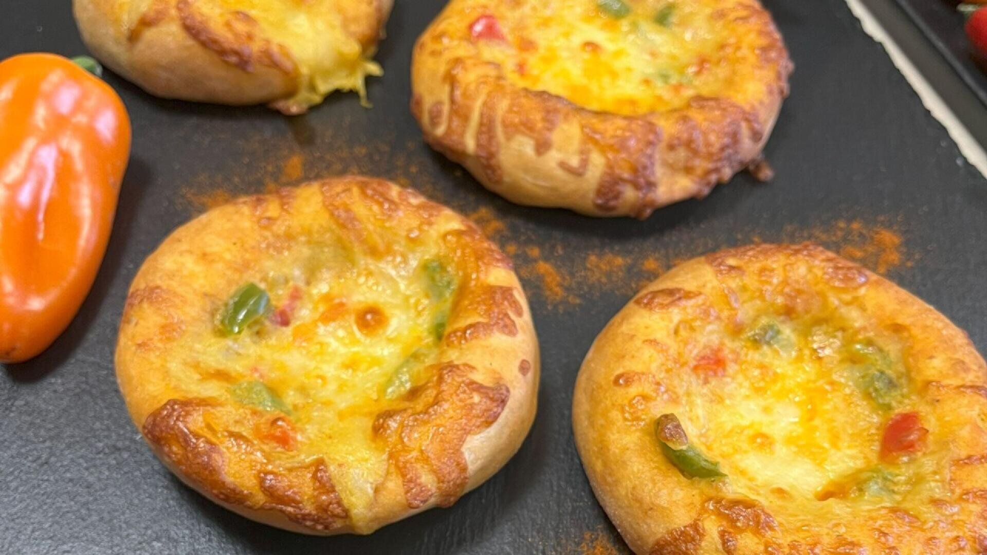Four mini cheese and vegetable pizzas with golden brown crusts are arranged on a black surface, next to an orange bell pepper. The pizzas have melted cheese and colorful vegetable pieces on top.