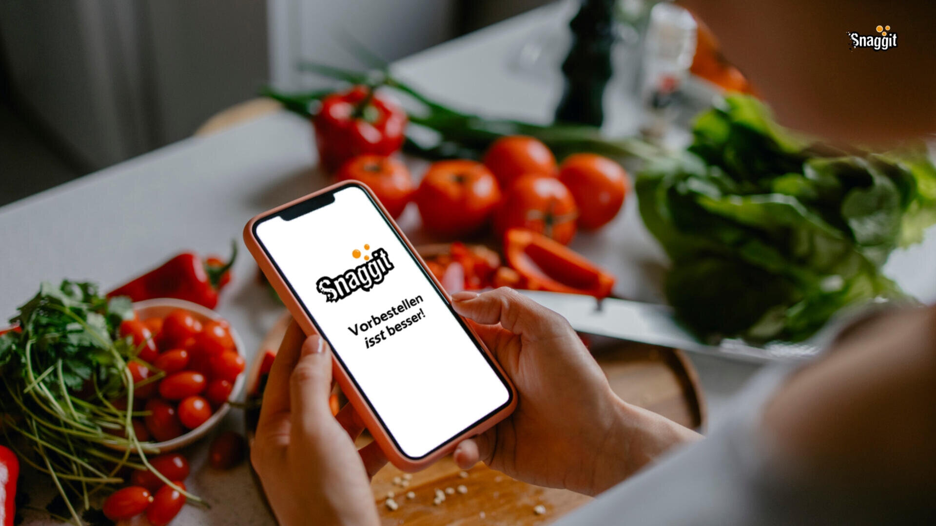 A person holds a smartphone displaying the Snaggit app in a kitchen, surrounded by fresh vegetables like tomatoes, lettuce, and herbs on a countertop.