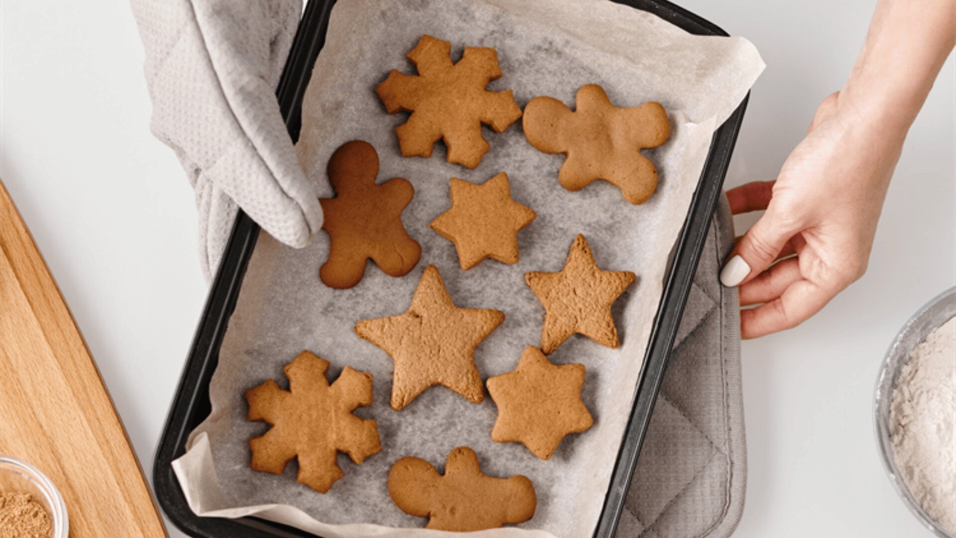 A person holds a baking tray lined with parchment paper, featuring freshly baked gingerbread cookies in star and gingerbread person shapes, resting on a quilted oven mitt.
