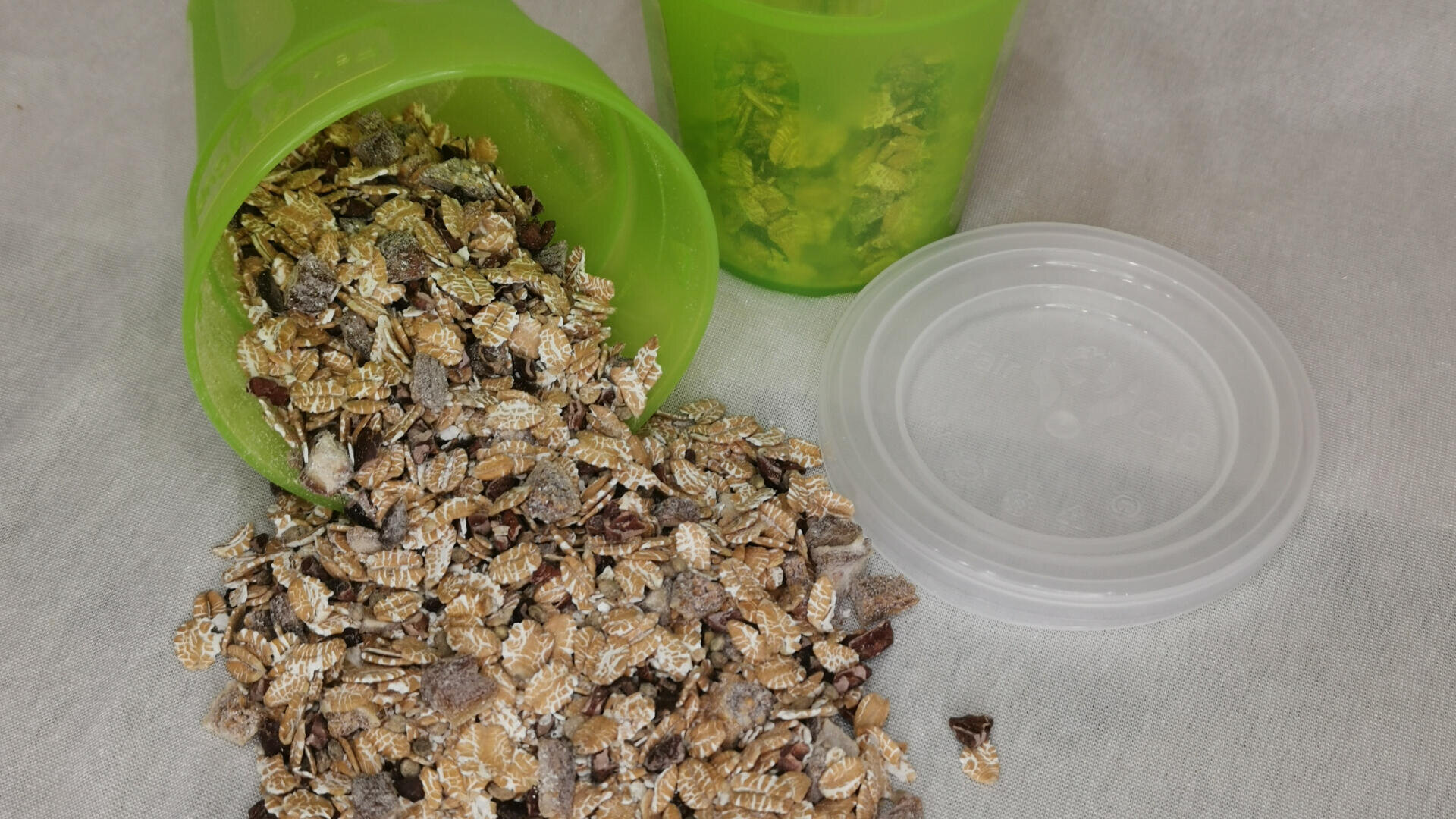 Ein grüner Plastikbecher ist umgekippt und verschüttet Müsli auf eine weiße Fläche. Ein weiterer identischer Becher, aufrecht stehend und teilweise mit Müsli gefüllt, steht in der Nähe, mit einem durchsichtigen Plastikdeckel daneben.