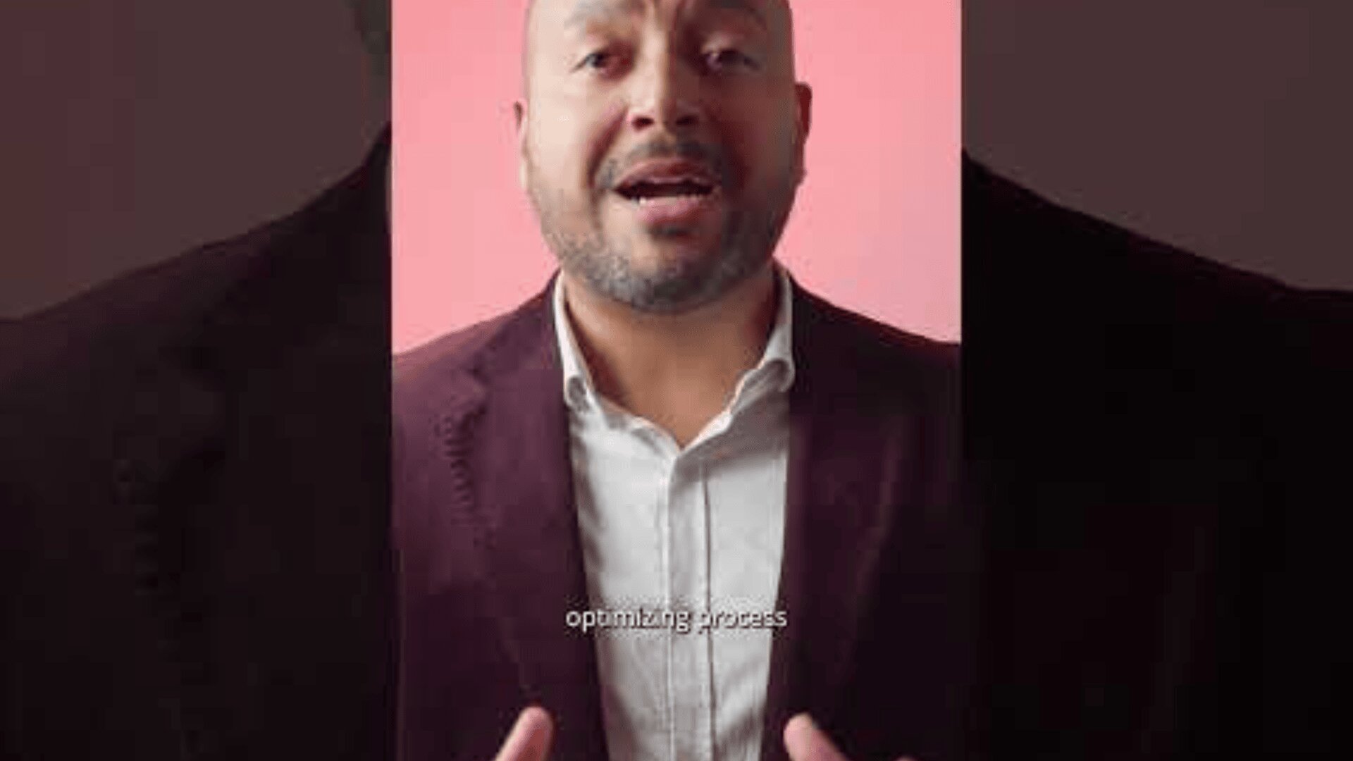 A man in a maroon blazer and white shirt speaks in front of a pink background, gesturing with his hands. Subtitles at the bottom read optimizing process.