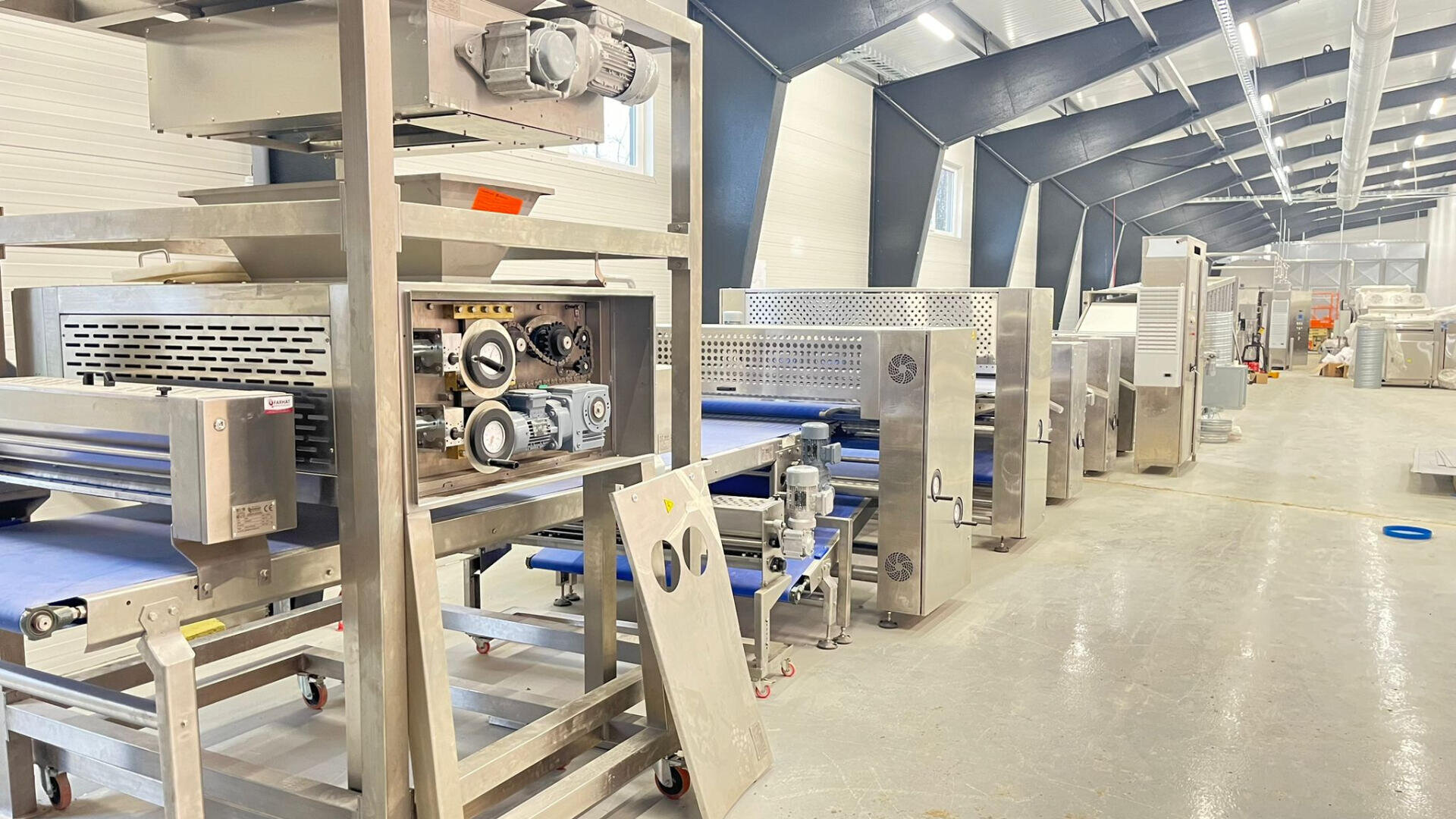 Rows of large industrial food processing machines made of stainless steel are lined up inside a brightly lit factory with a high, open ceiling. The floor is smooth and there are some blue conveyor belts visible.